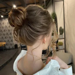 Upięcie włosów w kok na brązowych włosach z delikatnymi refleksami, widok z tyłu, złota biżuteria, biała koszula, neutralne tło z roślinami.