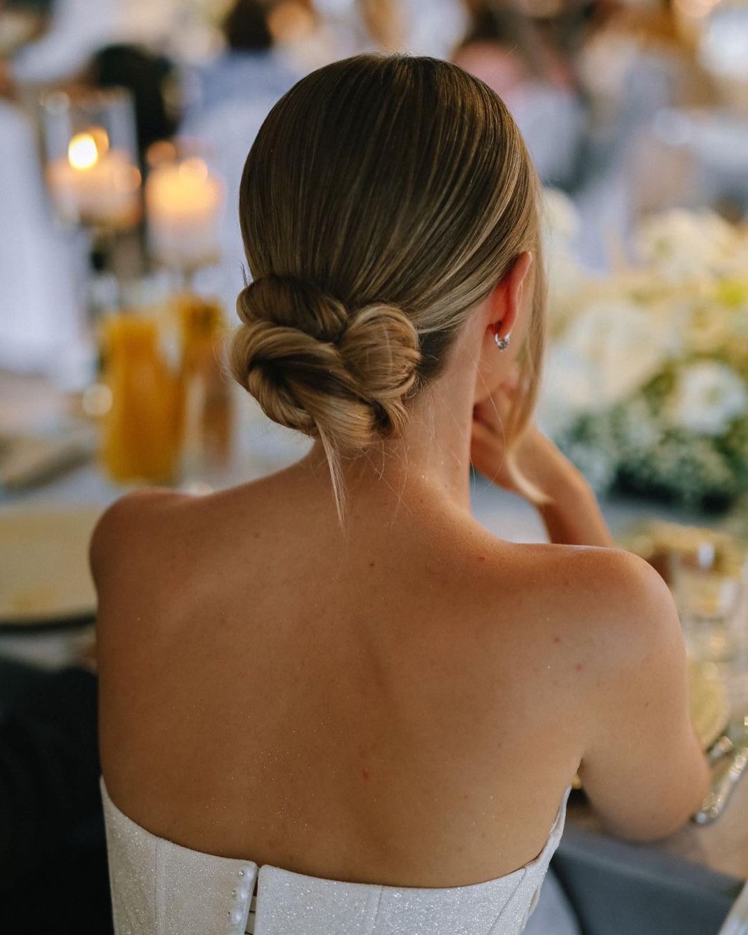 Elegancka upięcie włosów blond z pasemkami, widok z tyłu, delikatny makijaż, kolczyki-kółka, suknia na ramiączkach, stół z dekoracjami i napojami w tle.