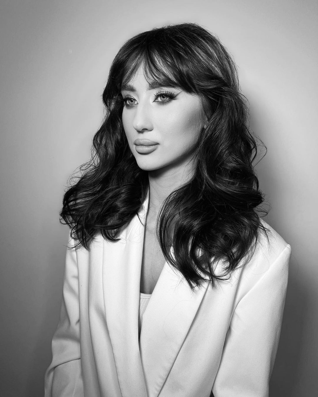 Portret kobiety z pełnym makijażem oczu i pofalowanymi, ciemnymi włosami z grzywką, ubrana w białą marynarkę, na jednolitym tle.