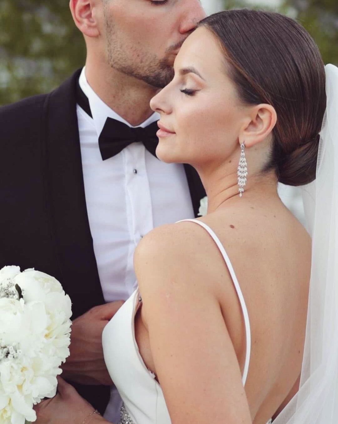 Panna młoda z upiętymi włosami i długimi, srebrnymi kolczykami, w białej sukni na ramiączkach, trzyma biały bukiet, a pan młody w czarnym smokingu całuje ją w czoło.