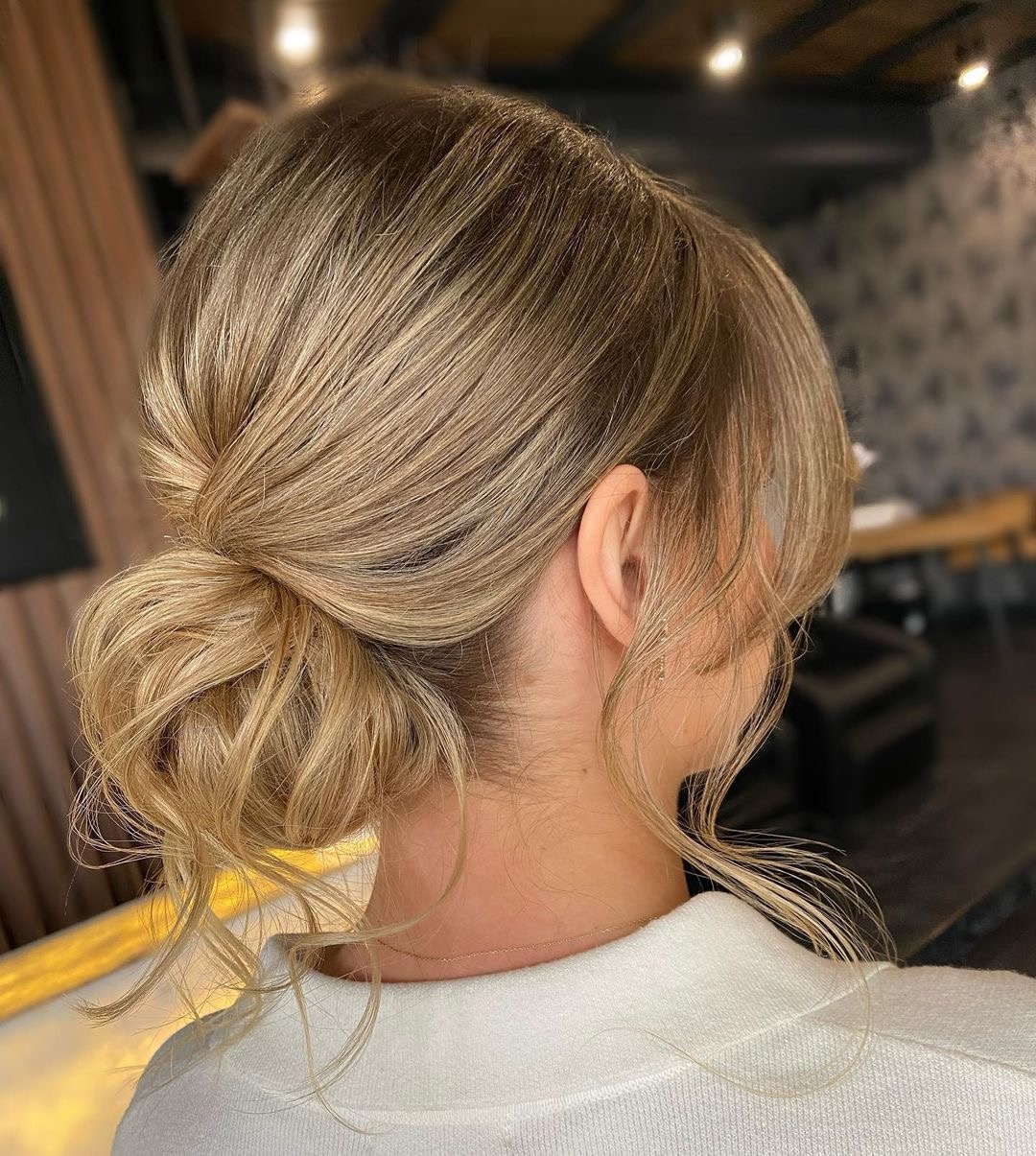 Upięcie włosów blond w niski kok z wypuszczonymi pasmami przy szyi i grzywką na bok, widok z tyłu na tle jasnego wnętrza salonu.