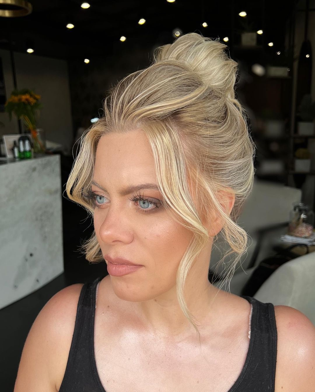 Blond włosy upięte w wysoki kok z wypuszczonymi, kręconymi pasmami przy twarzy, delikatny makijaż oczu i ust, widok z profilu w salonie.