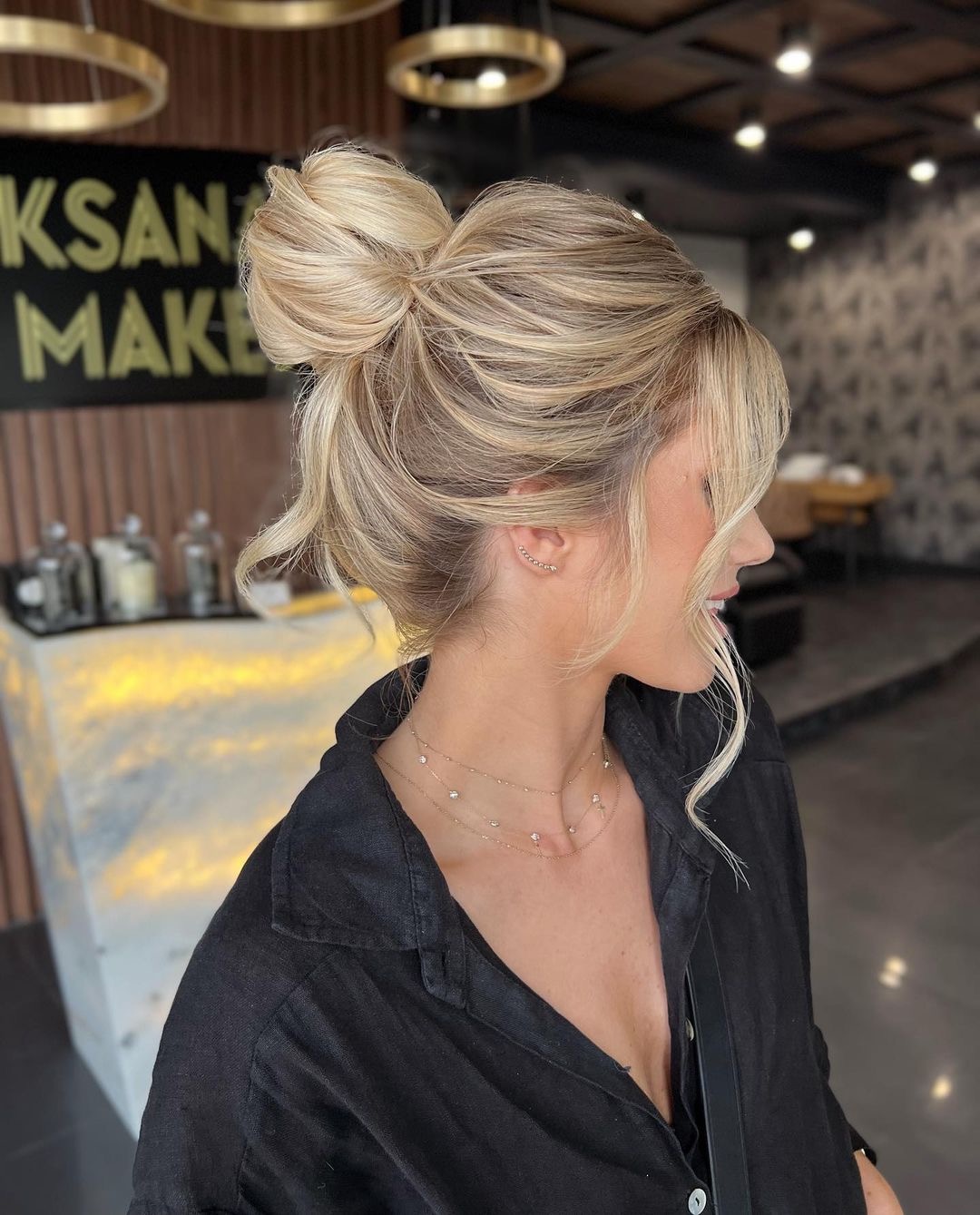 Profil kobiety z upięciem blond włosów w kok na czubku głowy, z delikatnymi kosmykami opadającymi na twarz, widoczne złote kolczyki i naszyjniki, czarna koszula.