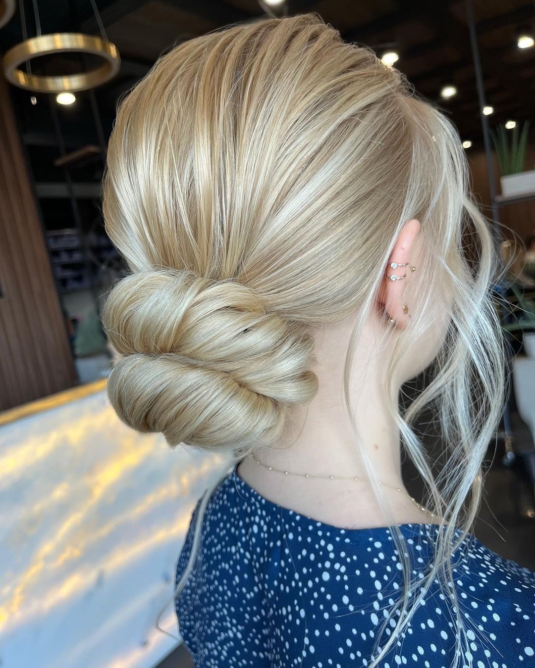 Blond włosy upięte w niski kok z boku głowy, widoczne kolczyki w uchu, niebieska bluzka w białe kropki.