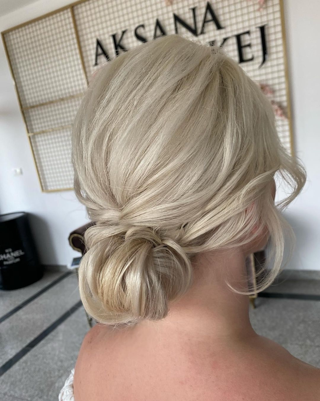 Upięcie blond włosów z tyłu głowy, kok w stylu messy bun, widoczny kark, w tle dekoracyjna siatka z napisem i flakon perfum.