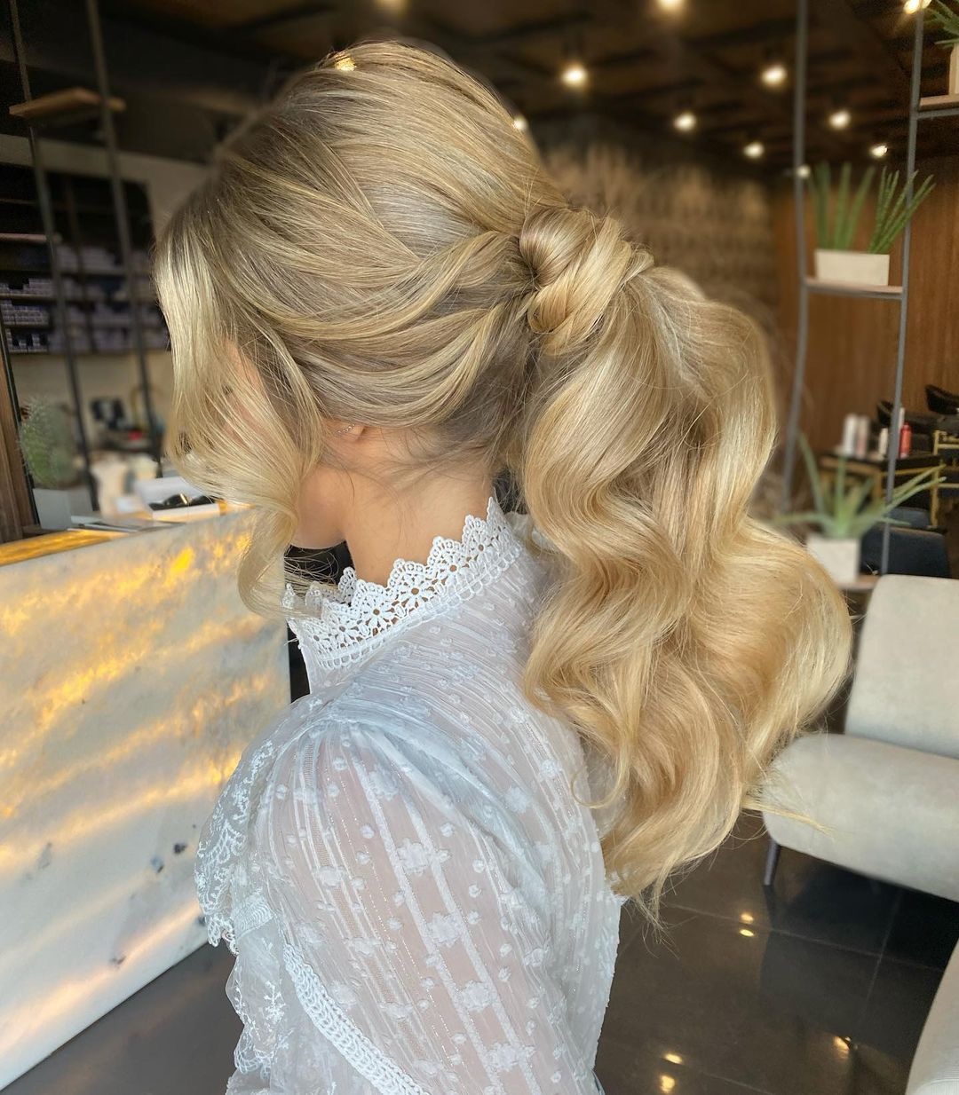 Blond włosy upięte w niski koński ogon z ozdobnym węzłem, delikatne fale, widok z boku, biała bluzka z koronkowym kołnierzykiem.