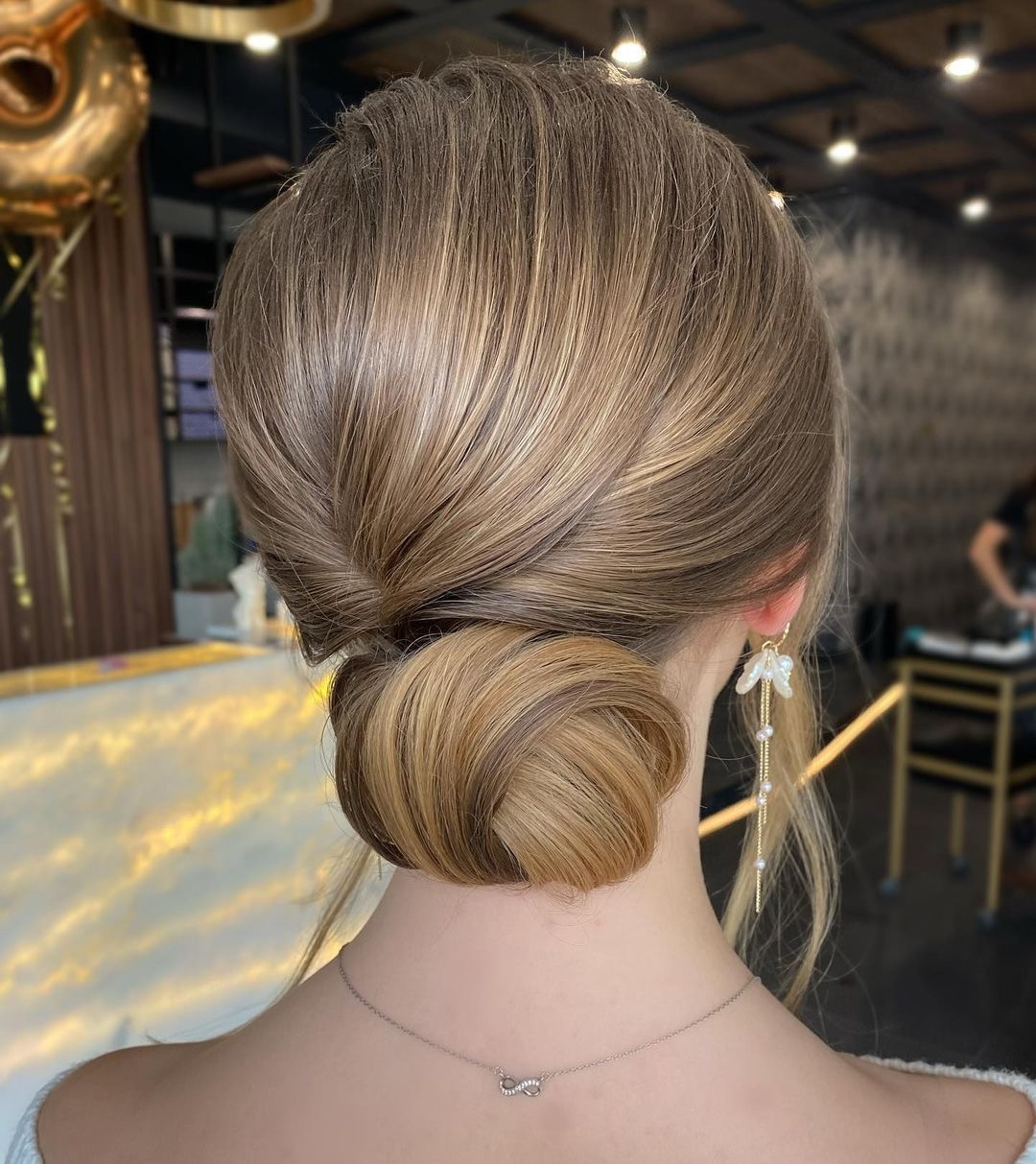 Elegancka fryzura upięta w niski kok z tyłu głowy, włosy blond z refleksami, widoczne długie kolczyki z perłami i delikatny srebrny naszyjnik.