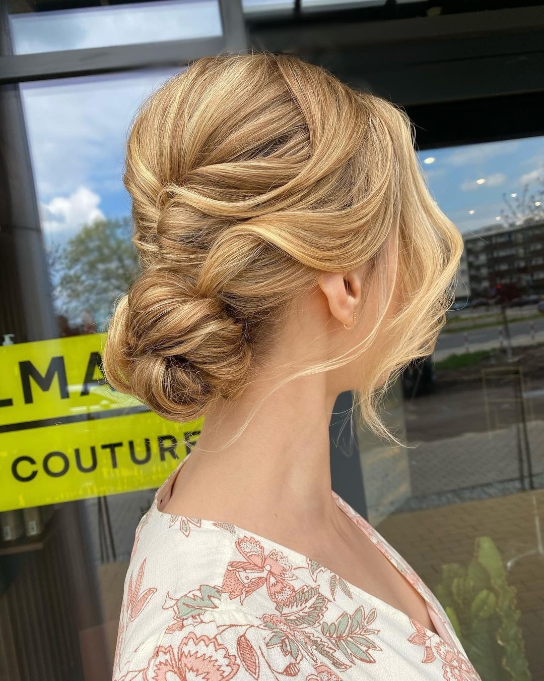 Eleganckie upięcie blond włosów z warkoczem i niskim kokiem, widok z profilu na tle szyby salonu fryzjerskiego.