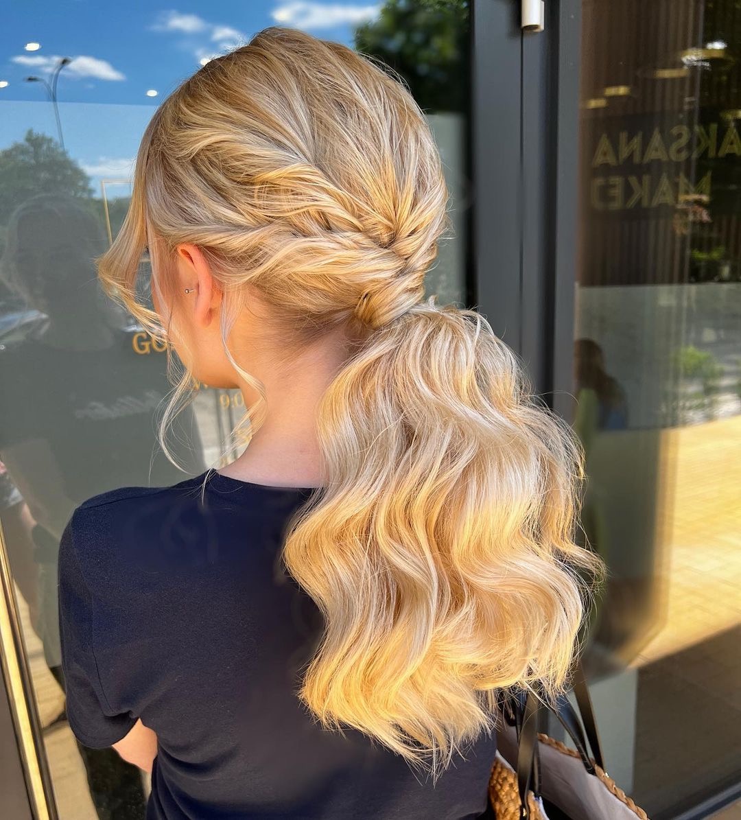 Upięcie blond włosów z zaplecionym warkoczem przechodzącym w niski kucyk z falowanymi końcówkami, widok z tyłu na tle szklanej witryny.