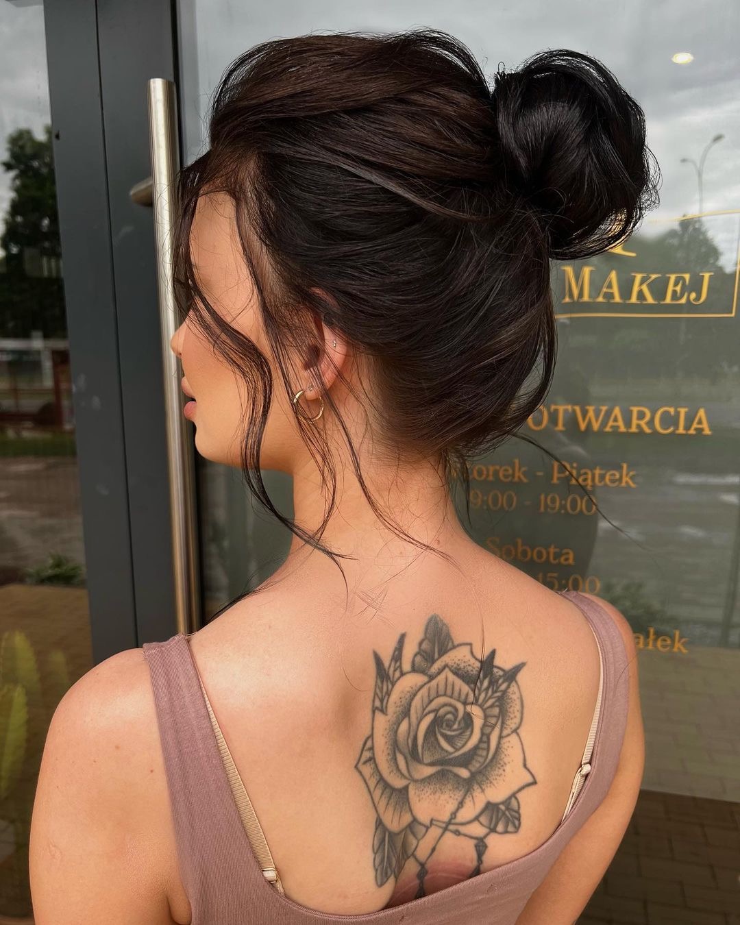 Upięcie włosów w kok na ciemnych włosach z pojedynczymi kosmykami opadającymi na twarz, widoczny tatuaż róży na plecach kobiety, na tle szyby z napisem 'Makej' i godzinami otwarcia.