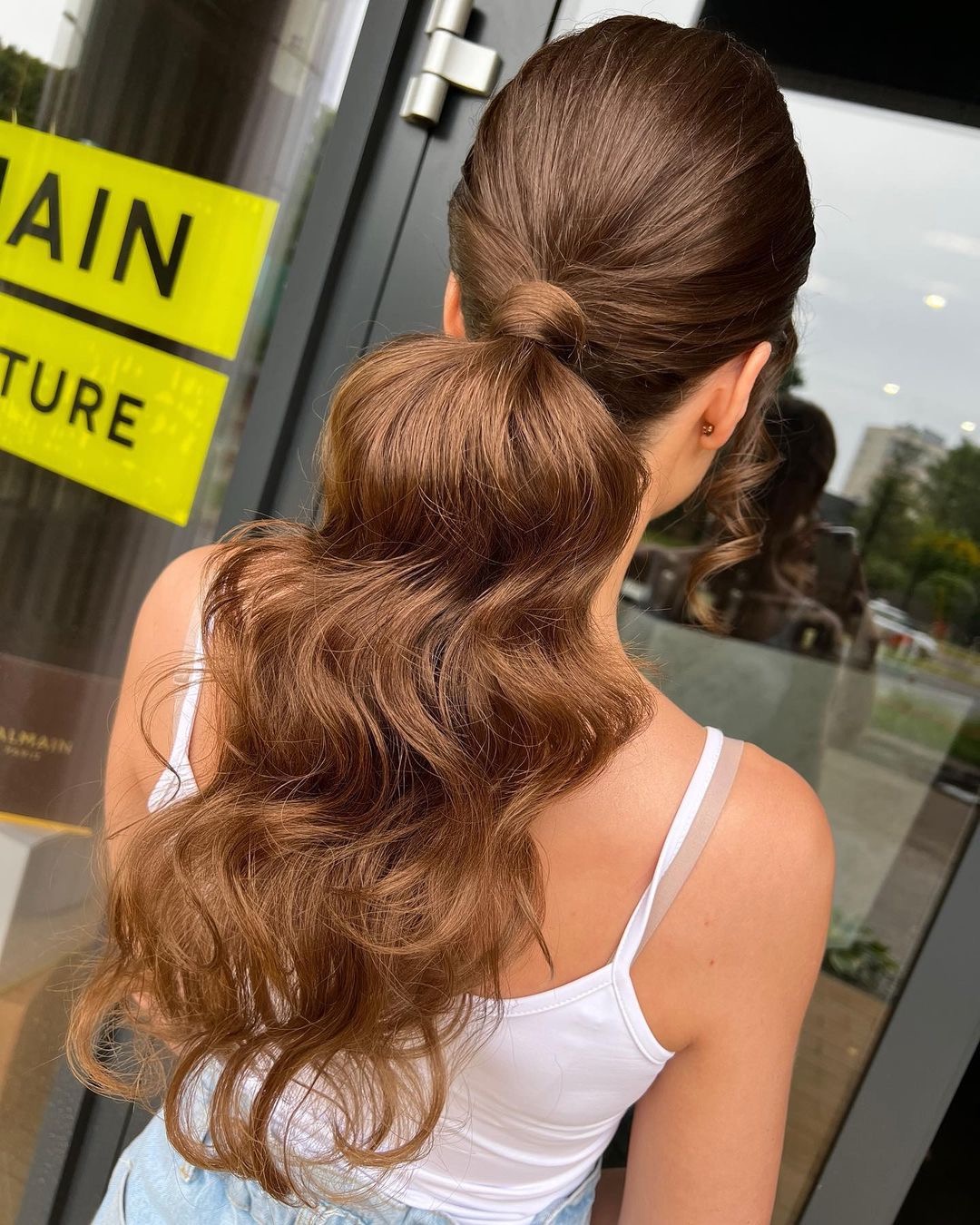 Upięte, brązowe włosy z falowanymi końcówkami, związane w wysoki kucyk z ukrytym wiązaniem, widok od tyłu na tle okna salonu.