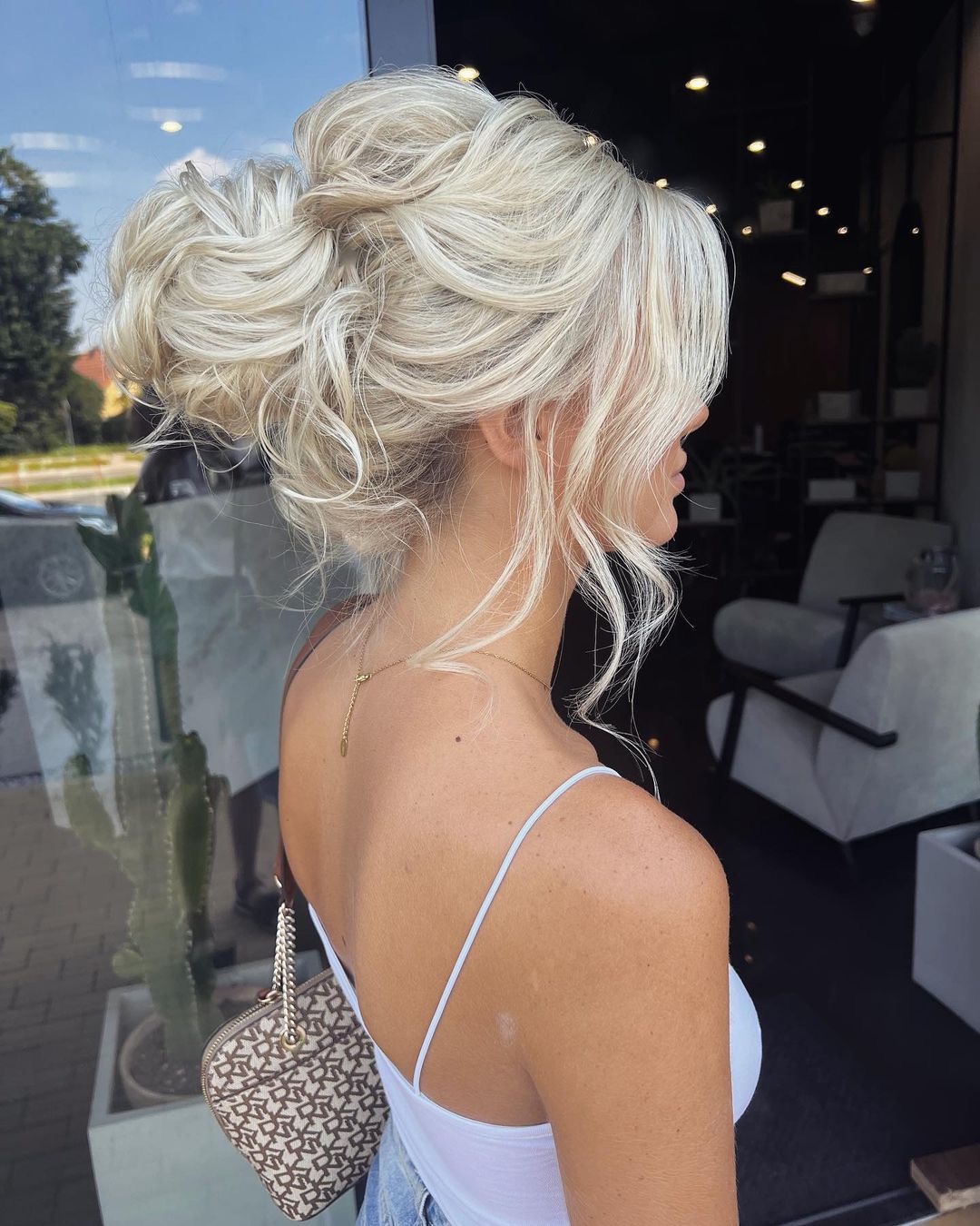 Upięcie blond włosów w dwa wysokie koki z wypuszczonymi pasmami, widok z profilu, delikatny makijaż, cienkie ramiączka białej bluzki, mała torebka na ramieniu