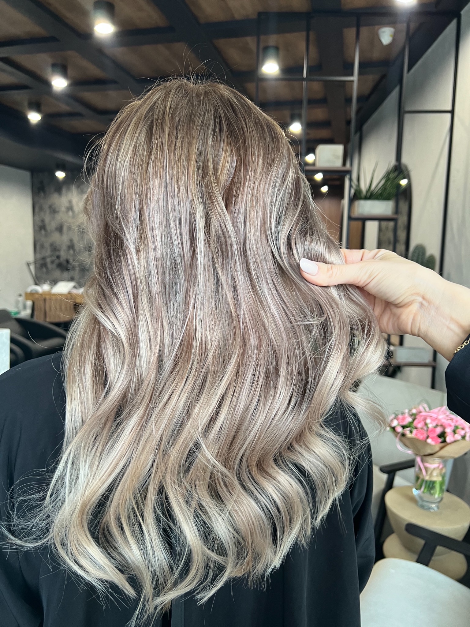 Włosy po koloryzacji sombre z chłodnymi refleksami, delikatne fale, dłoń z jasnym manicure dotyka kosmyków, w tle bukiet różowych kwiatów i elementy wystroju salonu.