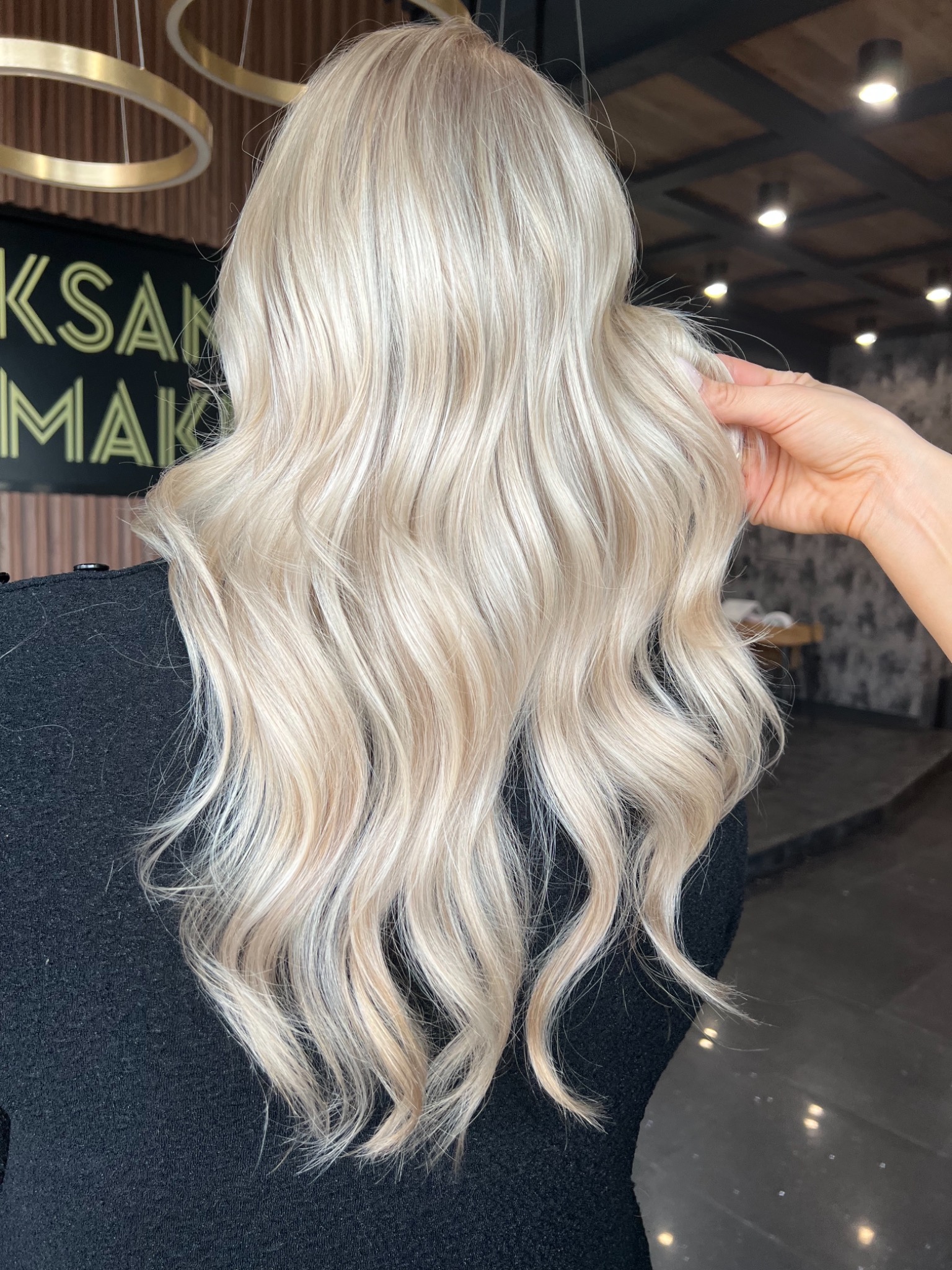 Tył głowy kobiety z długimi, platynowymi, falowanymi włosami z delikatnymi refleksami, układanymi przez dłoń w eleganckie fale w salonie fryzjerskim.