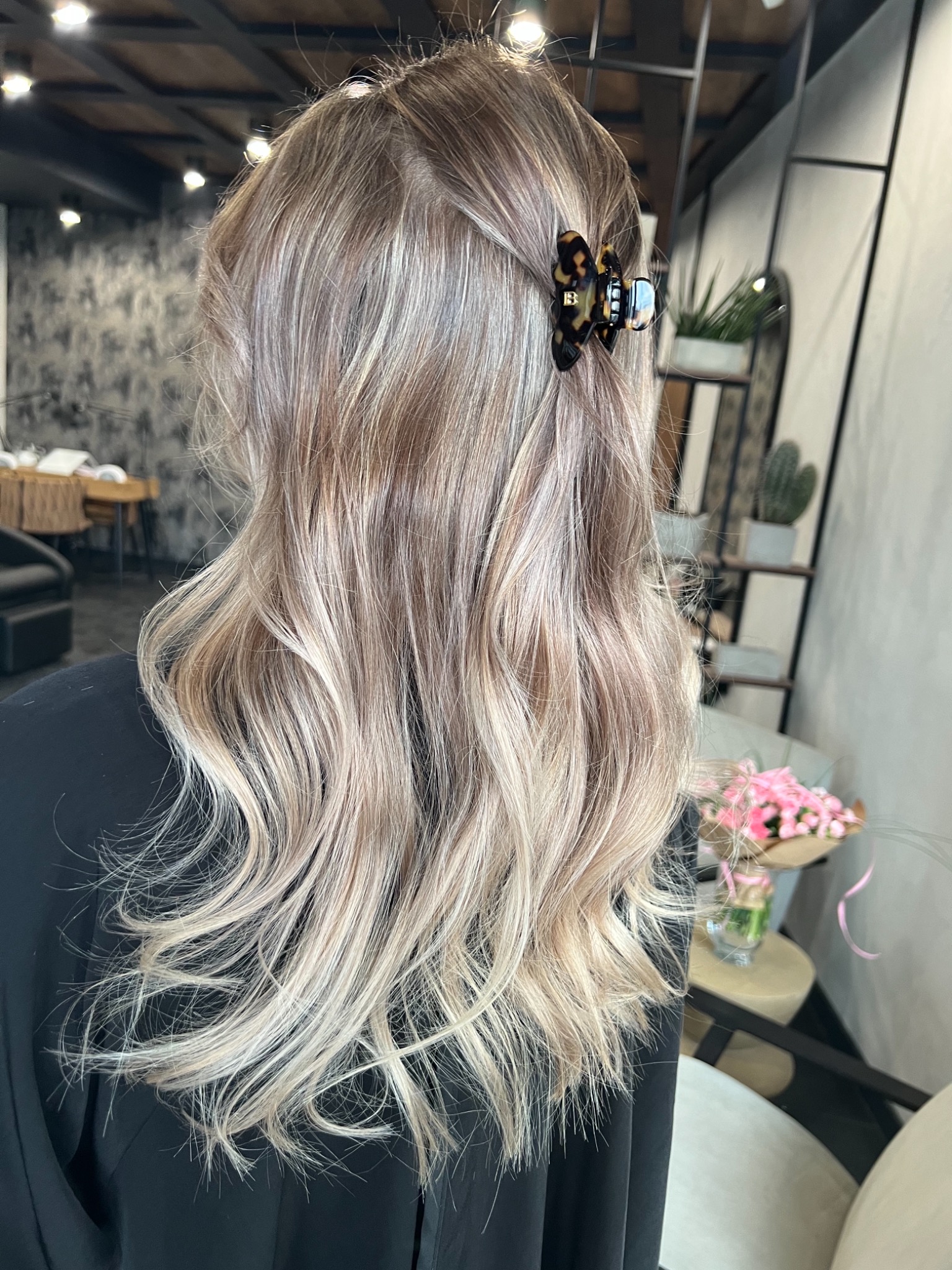 Długie, falowane włosy w odcieniach blondu z brązowym odrostem, upięte ozdobną spinką w kształcie motyla, widok z tyłu na tle wnętrza salonu.