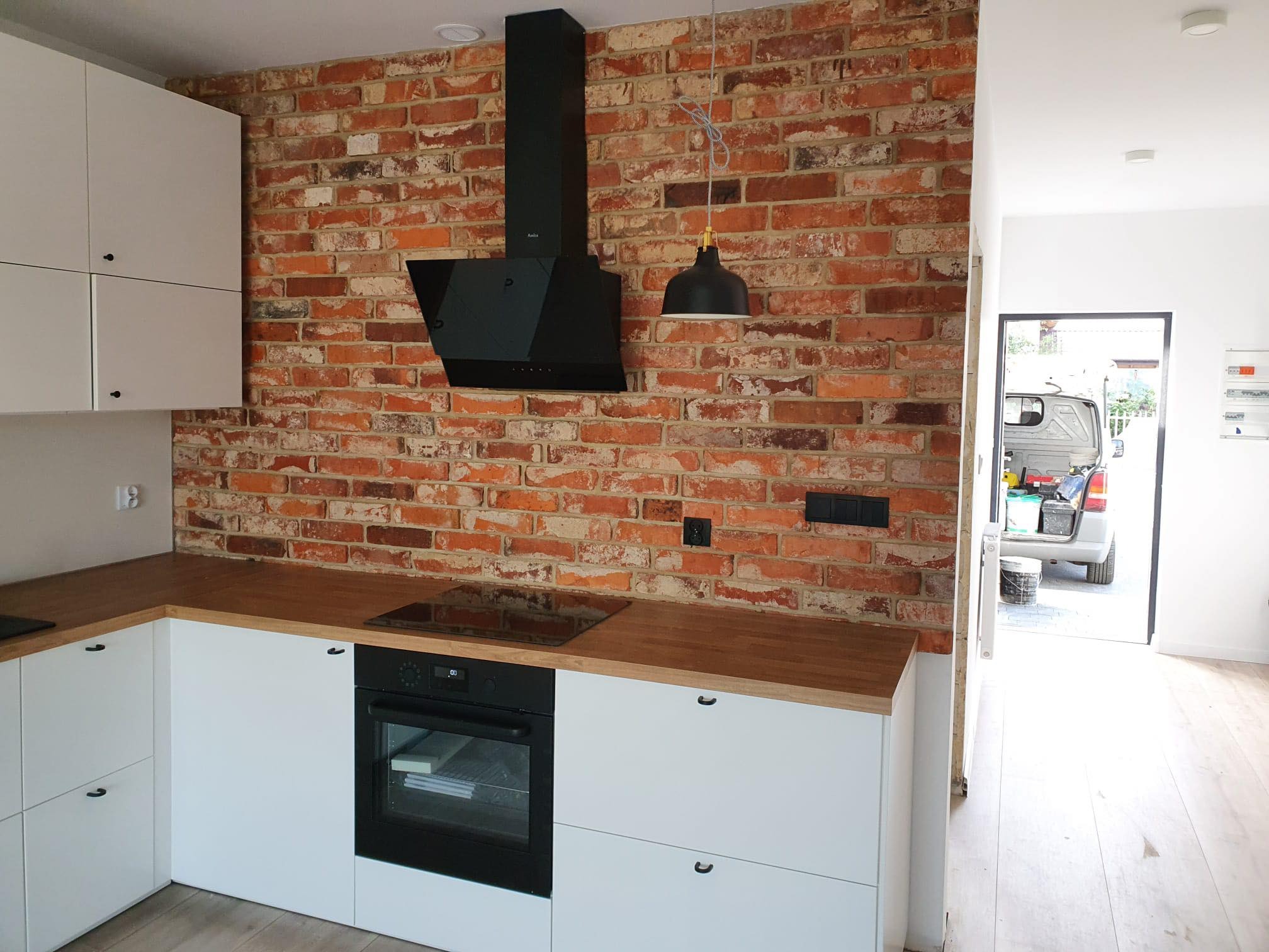 Kuchnia z ceglaną ścianą, białymi szafkami i czarnym okapem, widoczny fragment blatu z płytą indukcyjną i piekarnikiem, w tle otwarte drzwi i częściowo widoczny samochód dostawczy.