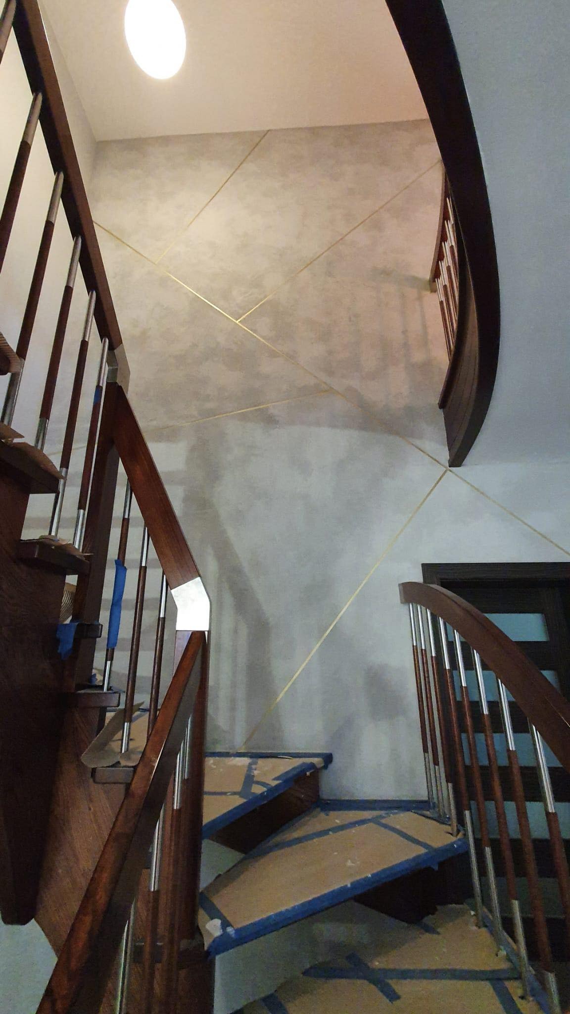 Schody drewniane z metalowymi balustradami zabezpieczone kartonem i taśmą malarską podczas prac wykończeniowych, na ścianie dekoracyjna szara farba z geometrycznymi złotymi liniami.