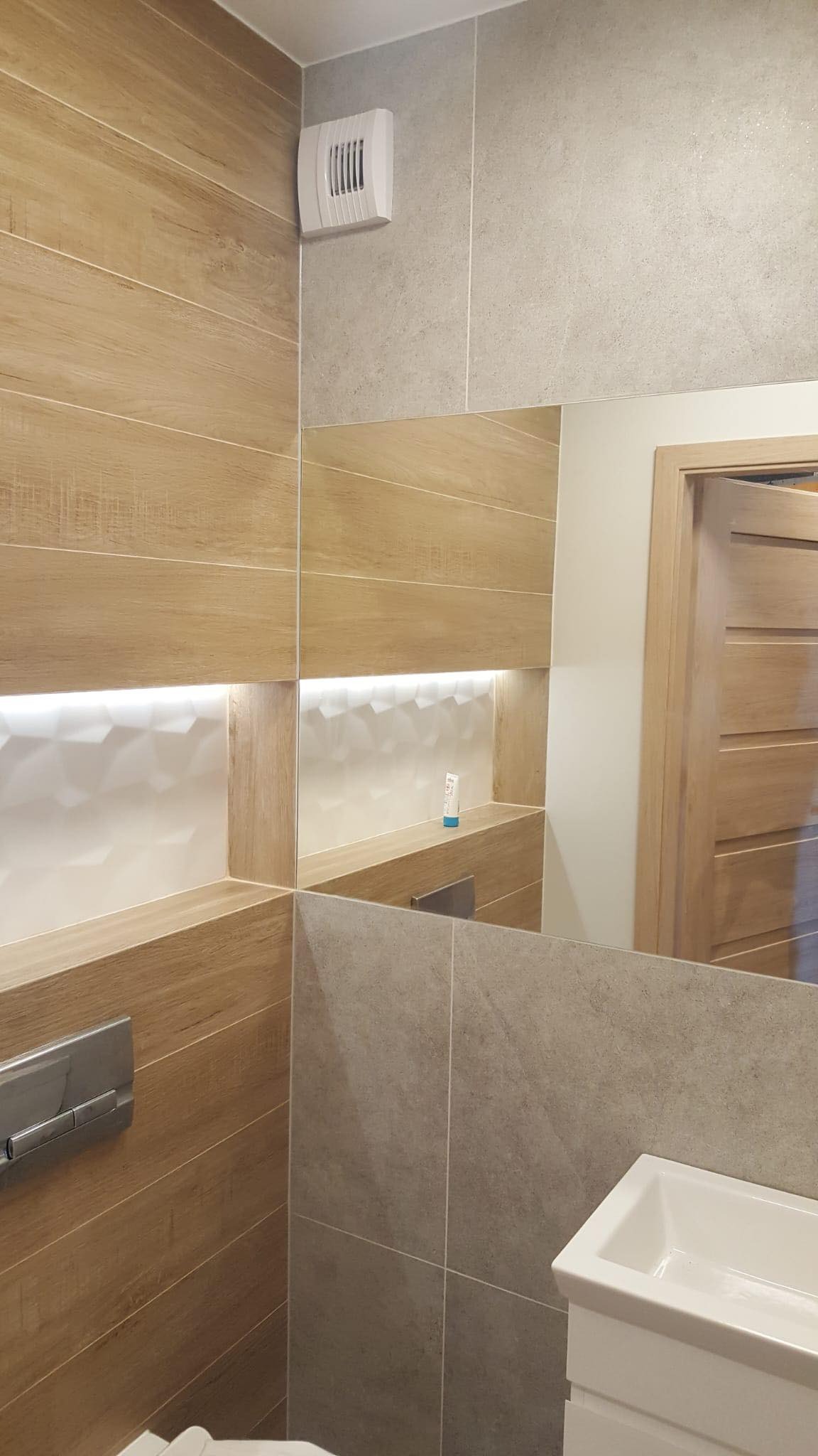 Nowoczesna łazienka z płytkami imitującymi drewno na ścianie, szarymi płytkami na pozostałej części ścian, podświetlaną wnęką z dekoracyjną białą ścianą 3D i minimalistyczną umywalką.