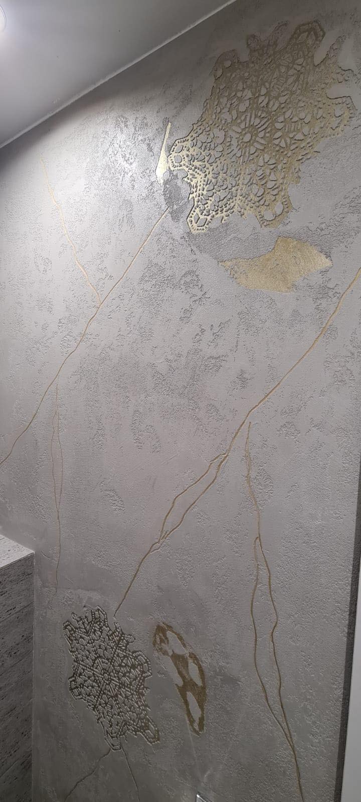Dekoracyjna ściana z szarą, teksturowaną farbą i złotymi, nieregularnymi wzorami przypominającymi koronki i żyły kamienia.