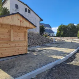 Nowa drewniana budka handlowa z czarnym dachem, ustawiona na betonowych fundamentach na placu budowy, z widokiem na budynek kościoła w tle.