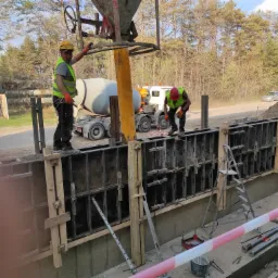 Wylewanie betonu do szalunków fundamentowych z betonomieszarki, dwóch pracowników w kaskach i kamizelkach odblaskowych nadzoruje proces, w tle widoczny las i przejeżdżający samochód.