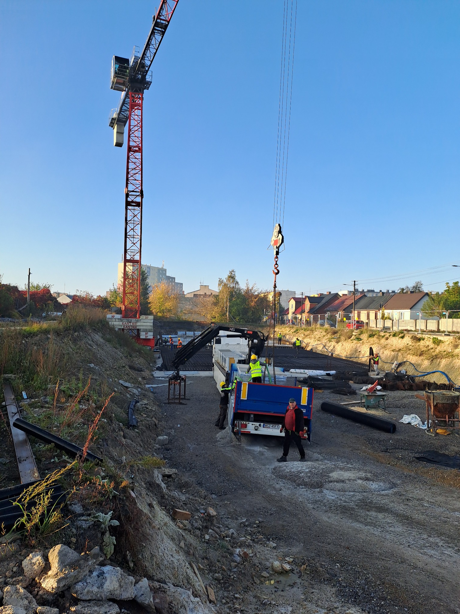 Widok na plac budowy z żurawiem wieżowym i pracownikami w kamizelkach odblaskowych, przygotowującymi zbrojenie fundamentów. W tle zabudowa jednorodzinna i blok mieszkalny.