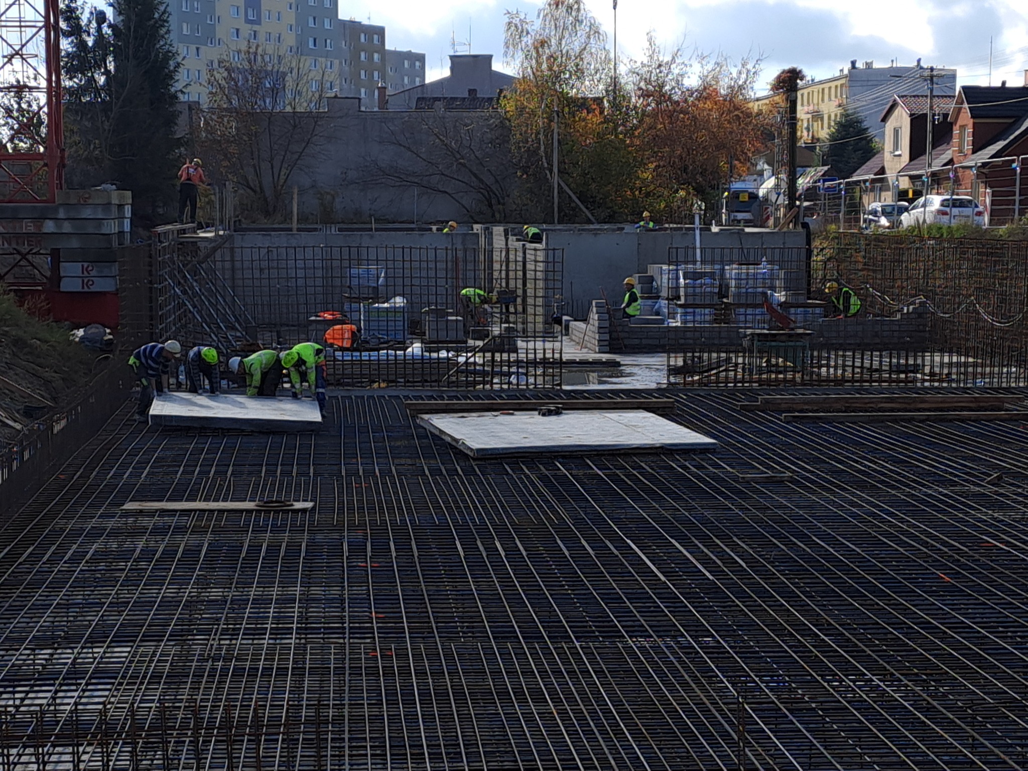 Siatka zbrojeniowa na budowie fundamentów, pracownicy w żółtych kamizelkach układają elementy szalunku, w tle budynki mieszkalne i dźwig.