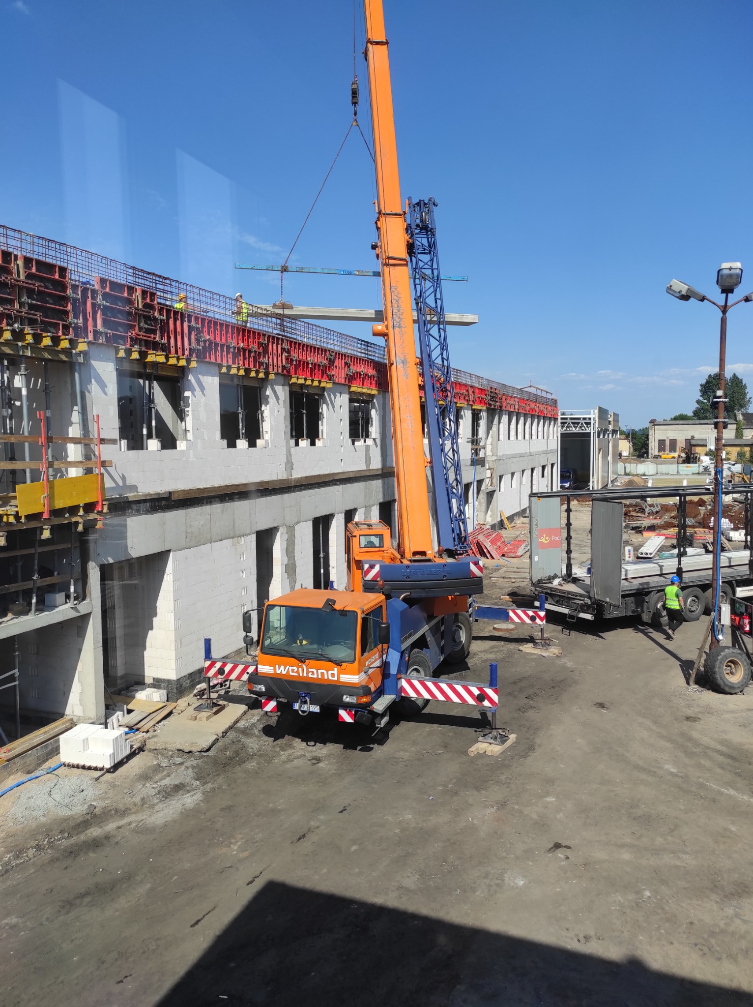 Budowa budynku z betonu komórkowego: pomarańczowy dźwig podnosi prefabrykat betonowy na placu budowy, widoczne szalunki i pracownicy w kaskach.