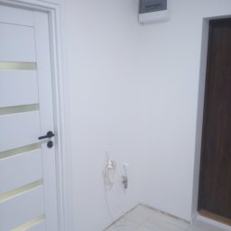 Dobre Wnętrze - Wnętrze po remoncie: białe ściany, jasna podłoga z płytek, białe drzwi z przeszkleniami, brązowe drzwi w tle, wystająca instalacja wodna i skrzynka elektryczna na ścianie.