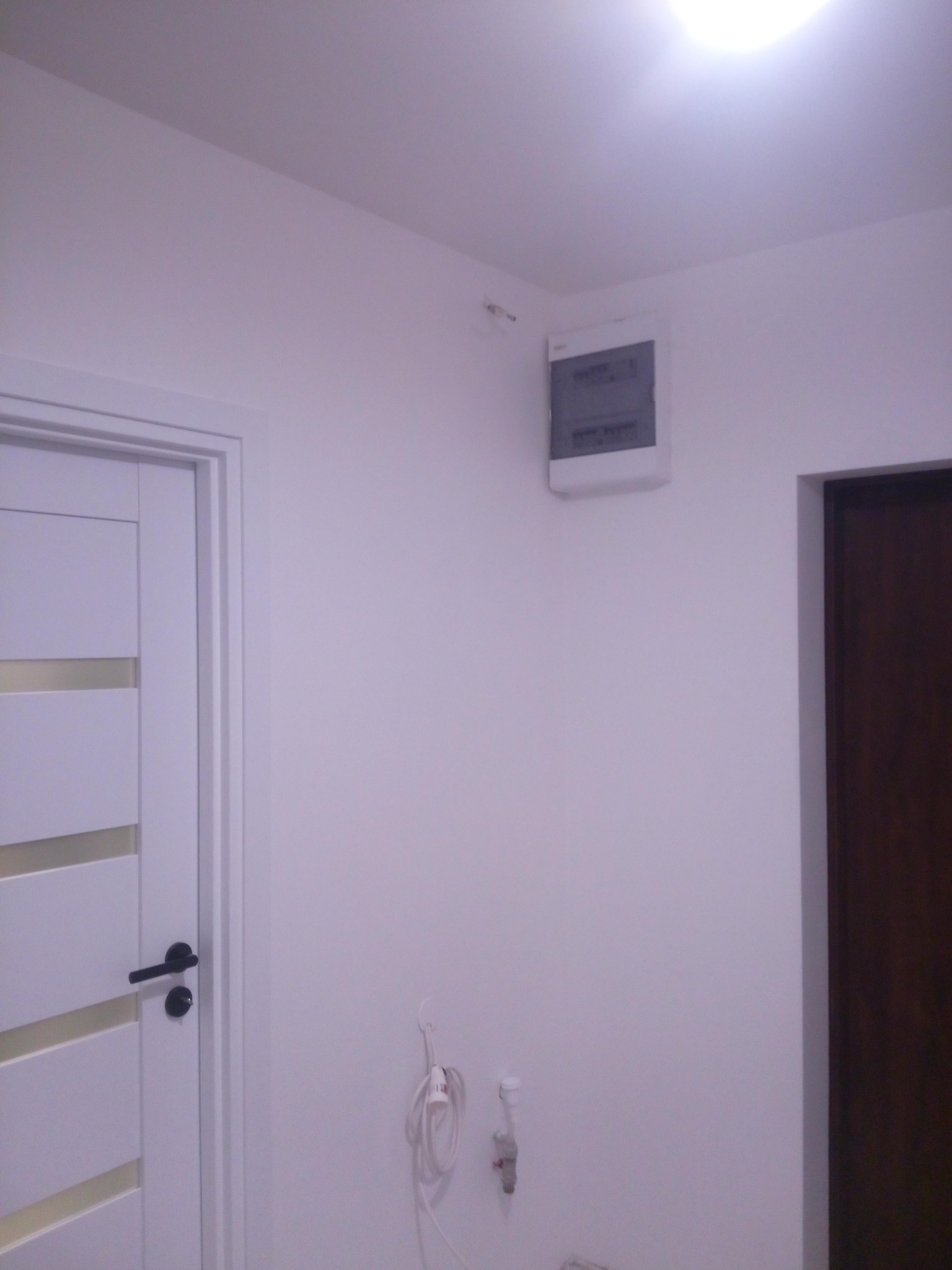 Wnętrze po odświeżeniu: białe ściany, nowa biała drzwi z poziomymi przeszkleniami i czarną klamką, skrzynka elektryczna na ścianie, gniazdko z przewodem i kranik.