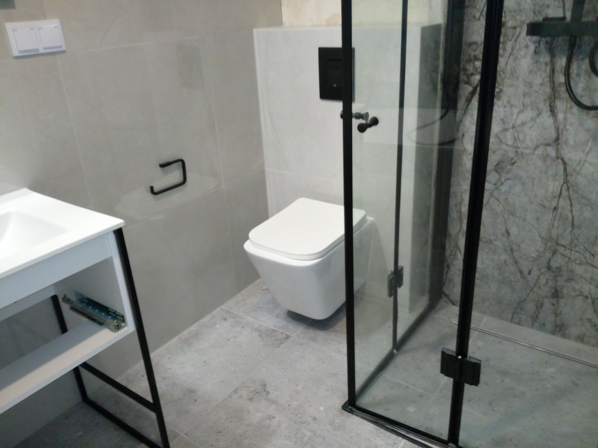 Nowoczesna łazienka z białą wiszącą toaletą, umywalką na czarnym stelażu i kabiną prysznicową z czarnymi profilami, wykończona szarymi płytkami imitującymi kamień.