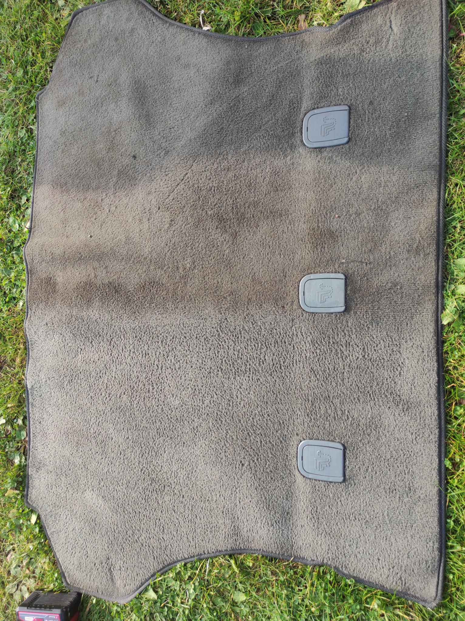 Brudny, szary dywanik samochodowy z trzema plastikowymi zatrzaskami leżący na trawie, widoczne ślady użytkowania i plamy.