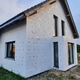 Niedokończony budynek jednorodzinny z białym styropianem na elewacji, widoczne czarne ramy okienne i elementy konstrukcji dachu, teren wokół budynku niezagospodarowany.