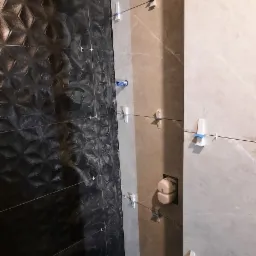 Układanie płytek w łazience: czarne płytki z wypukłym wzorem na jednej ścianie, jasnoszare na drugiej, widoczne krzyżyki dystansowe i poziomowanie laserowe, nieukończona podłoga.