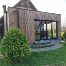 Nowoczesna dobudówka do domu z elewacją z pionowych desek, dużymi oknami i kamiennymi schodami wejściowymi.