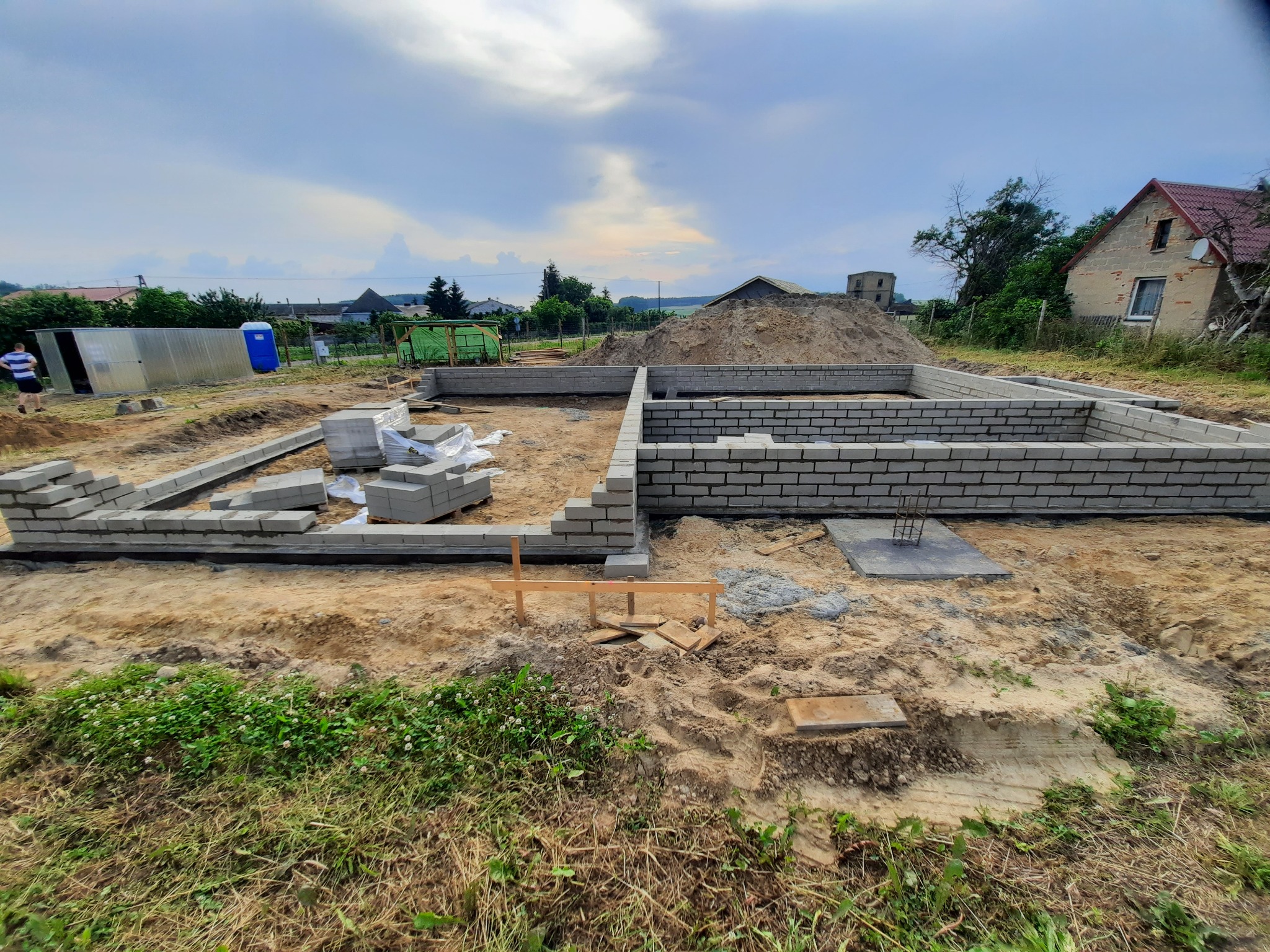 Budowa fundamentów domu z bloczków betonowych, widoczne pierwsze warstwy muru, przygotowane bloczki, teren budowy z piaskiem i trawą, w tle budynki gospodarcze i dom jednorodzinny.