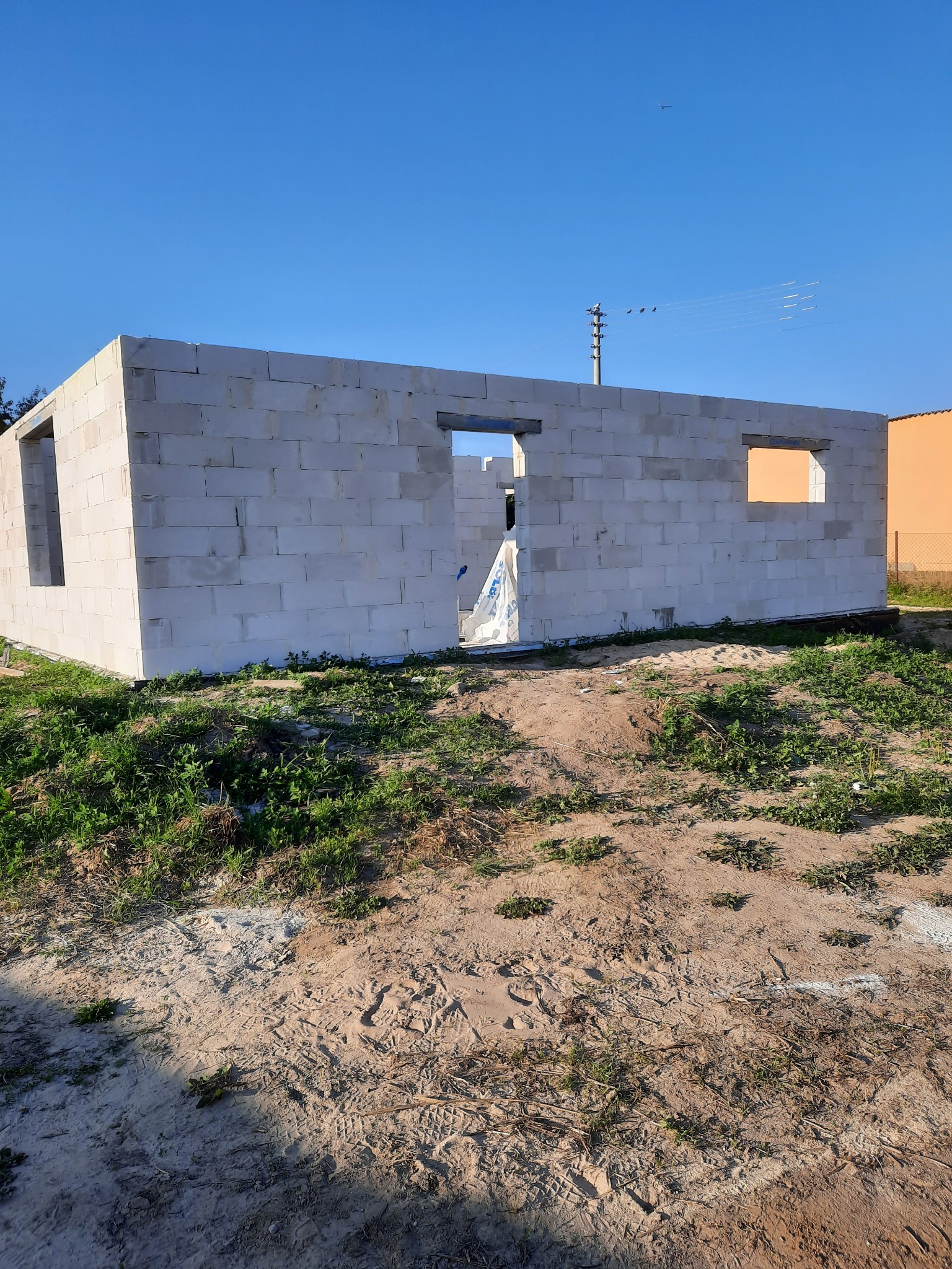 Surowy stan ścian parterowego domu z betonu komórkowego w trakcie budowy, widoczne otwory okienne i drzwiowe, teren wokół niezagospodarowany.