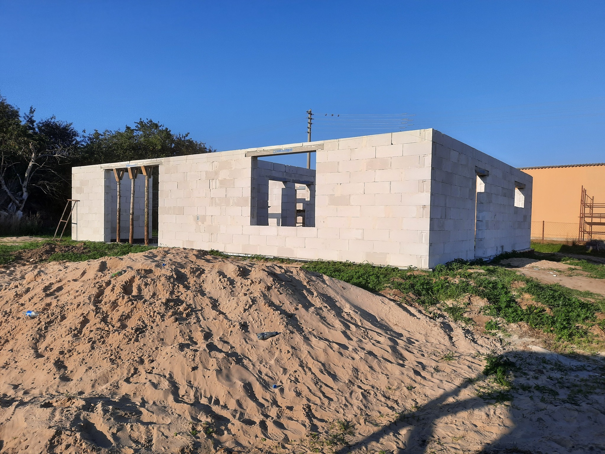 Surowy stan domu z bloczków, z widocznymi otworami okiennymi i drzwiowymi, podparty drewnianymi stemplami, na tle błękitnego nieba i sterty piasku.