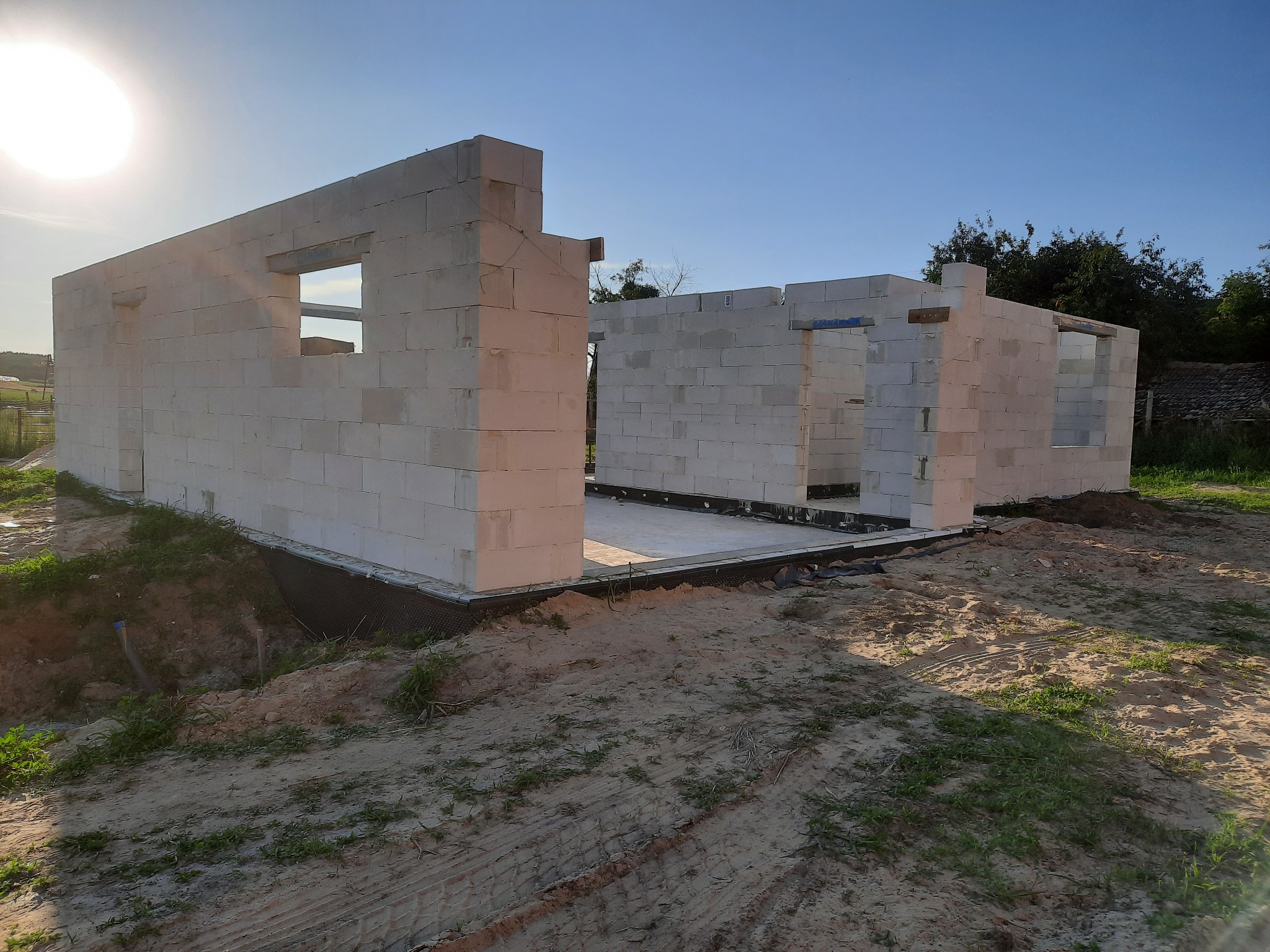 Wznoszenie ścian parterowego domu murowanego z betonu komórkowego, widoczne otwory okienne i drzwiowe, teren budowy z piaskiem i zielenią w tle.