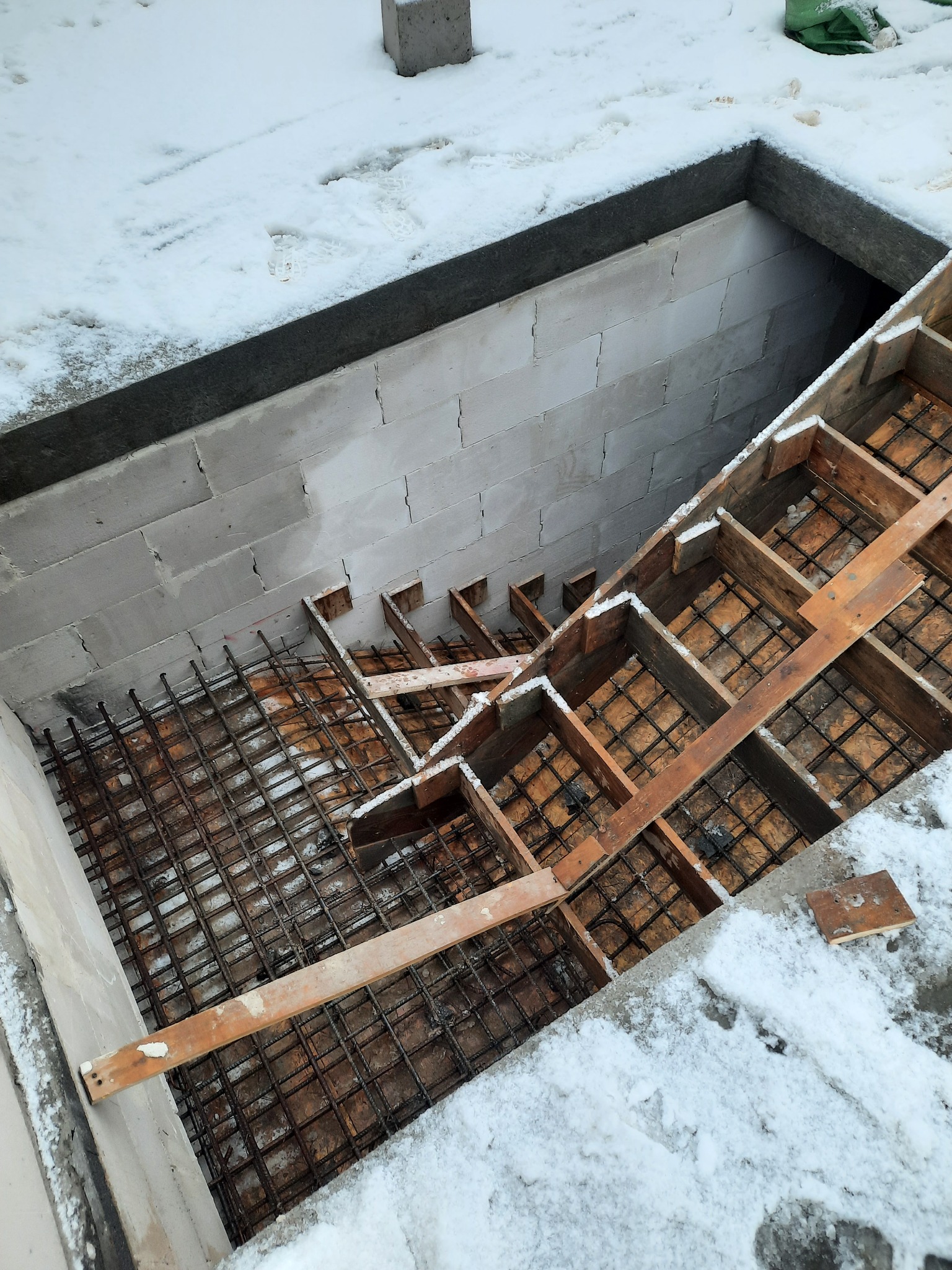 Fundament domu w trakcie budowy: zbrojenie stalowe w drewnianym szalunku, ściany z bloczków betonowych, lekko przysypane śniegiem.
