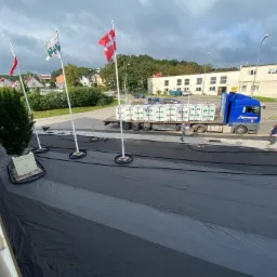 Ciężarówka z naczepą załadowana paletami z materiałami budowlanymi, stojąca na czarnej folii rozłożonej na ziemi, w tle budynek i maszty z flagami.