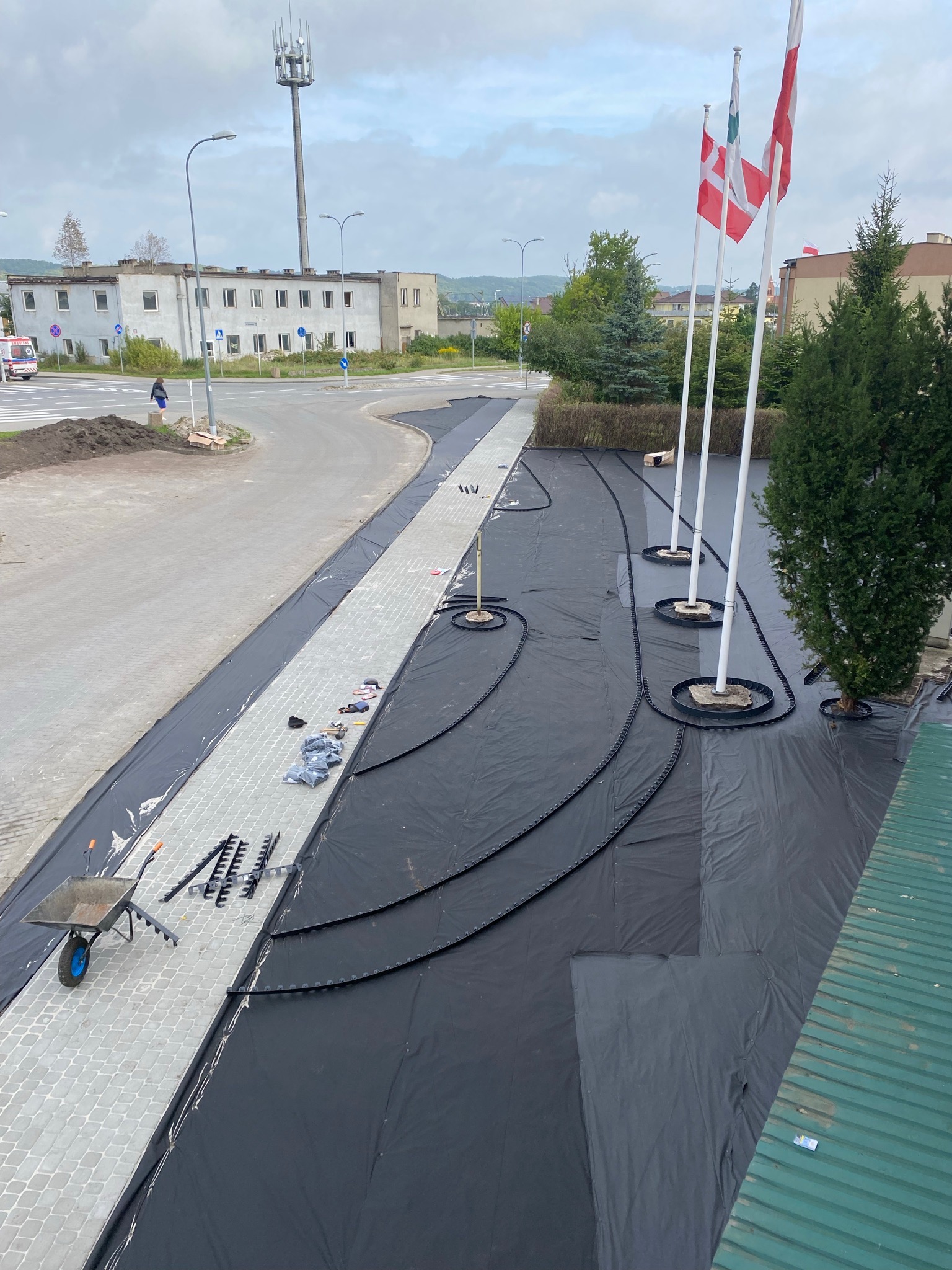 Przygotowanie terenu pod nasadzenia w przestrzeni miejskiej: rozłożona czarna agrowłóknina, system nawadniania wokół masztów flagowych, narzędzia ogrodnicze i taczka na kostce brukowej.