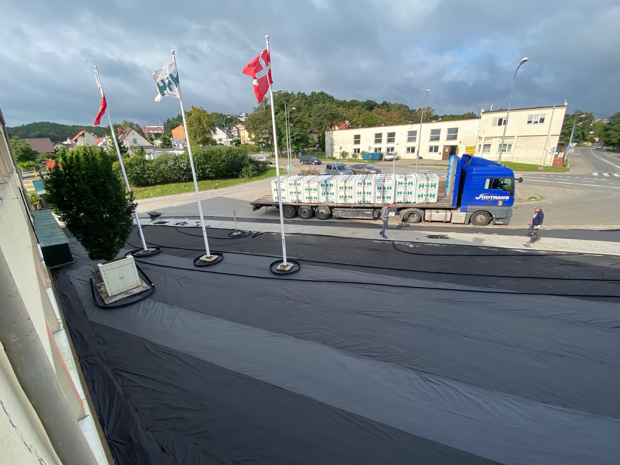 Ciężarówka z naczepą załadowana paletami z materiałami budowlanymi, stojąca na czarnej folii rozłożonej na ziemi, w tle budynek i maszty z flagami.