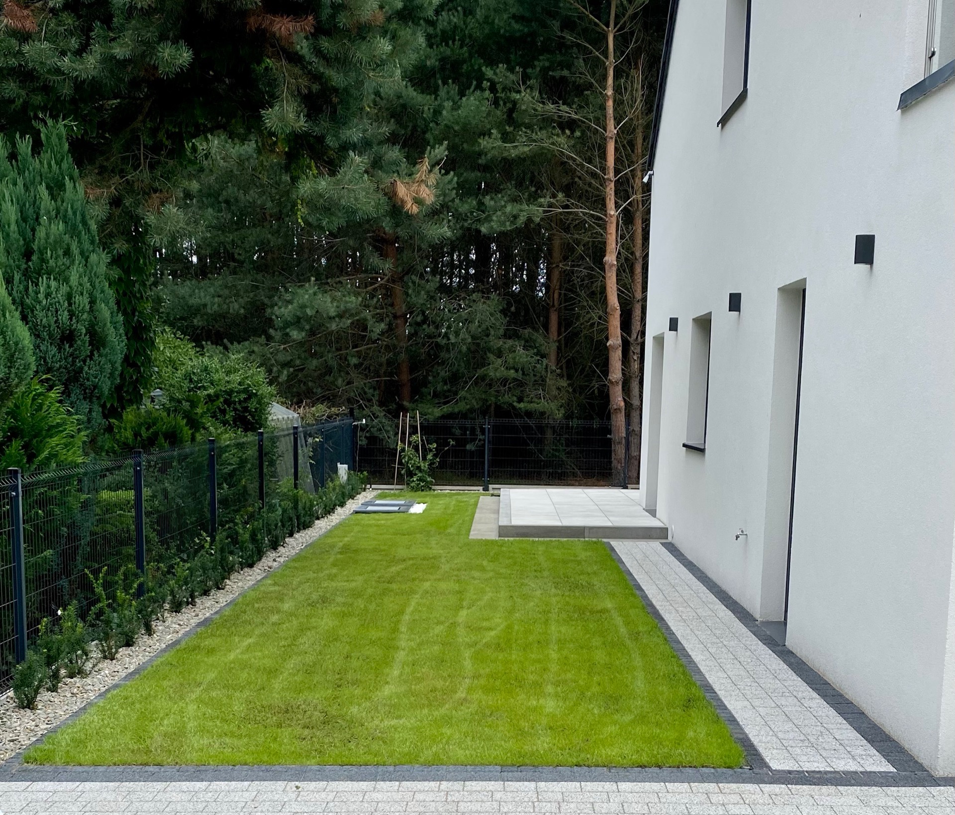 Świeżo skoszony trawnik przylegający do nowoczesnego domu z białymi ścianami, obrzeżem z kostki granitowej i podwyższonym tarasem z płyt, otoczony zielenią i ogrodzeniem panelowym.