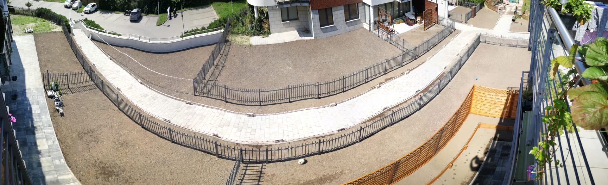 Widok z góry na nowo zaprojektowany ogród z krętą ścieżką z jasnej kostki brukowej, obramowaną niskim, ciemnym ogrodzeniem metalowym, otoczony świeżo przygotowaną ziemią pod trawnik, z fragmentami...