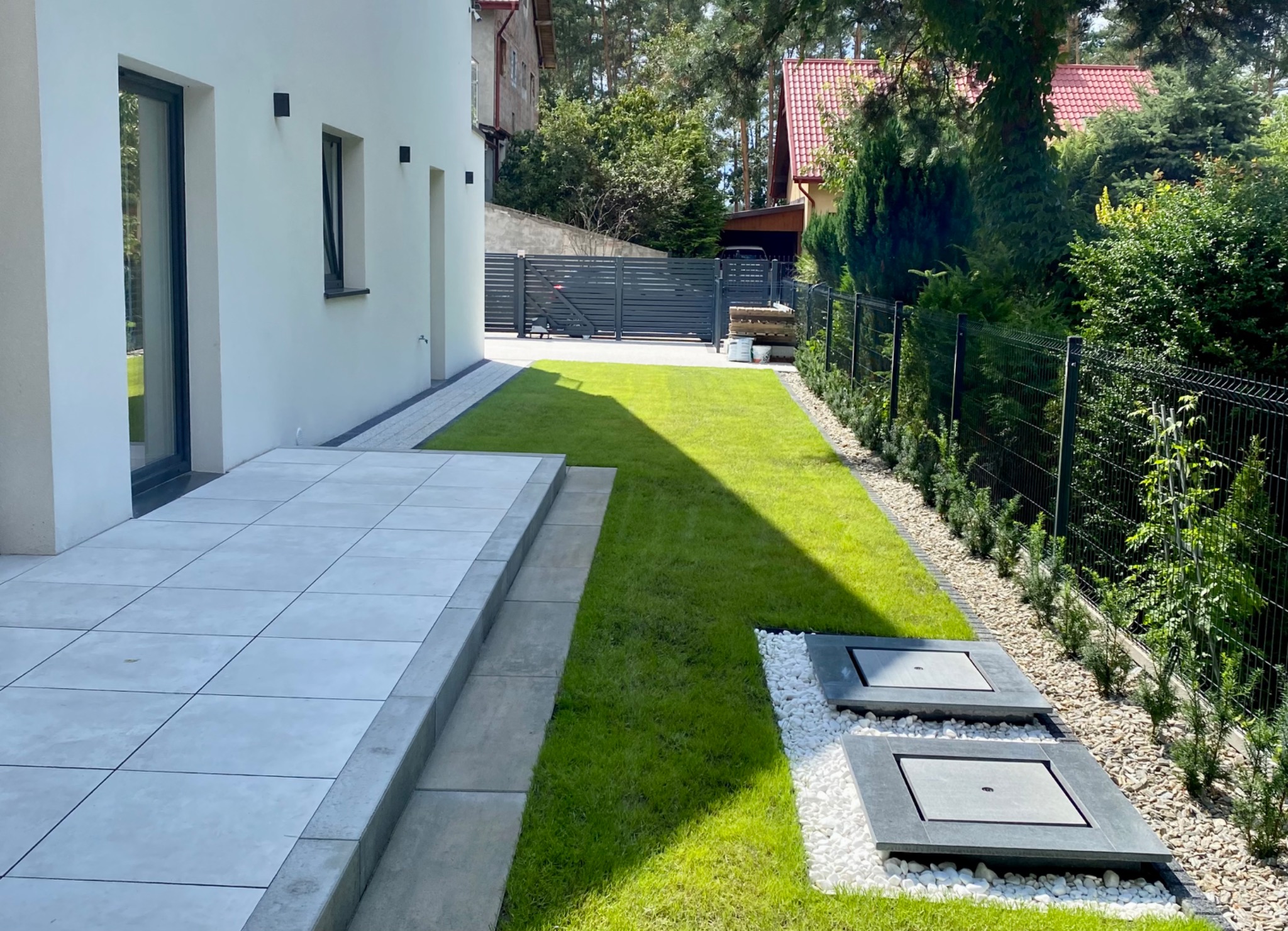 Nowoczesny ogród z równo przystrzyżonym trawnikiem, szarą kostką brukową, białymi kamieniami dekoracyjnymi, ciemną, metalową siatką ogrodzeniową i minimalistycznym domem w tle.