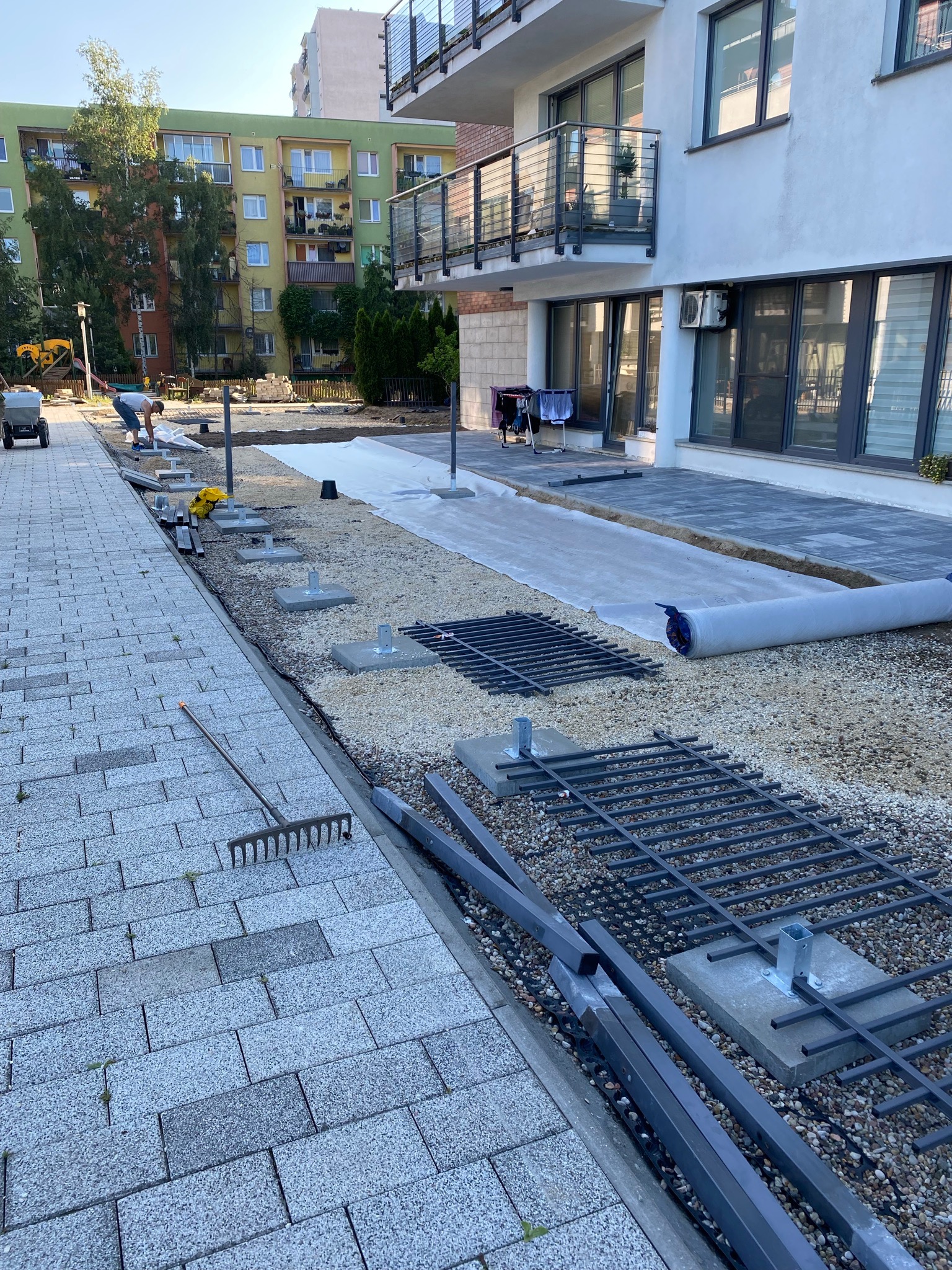 Przygotowanie terenu pod ogród: rozłożona geowłóknina, ustawione betonowe podstawy pod konstrukcję, widoczny mężczyzna przy pracy oraz narzędzia ogrodnicze.