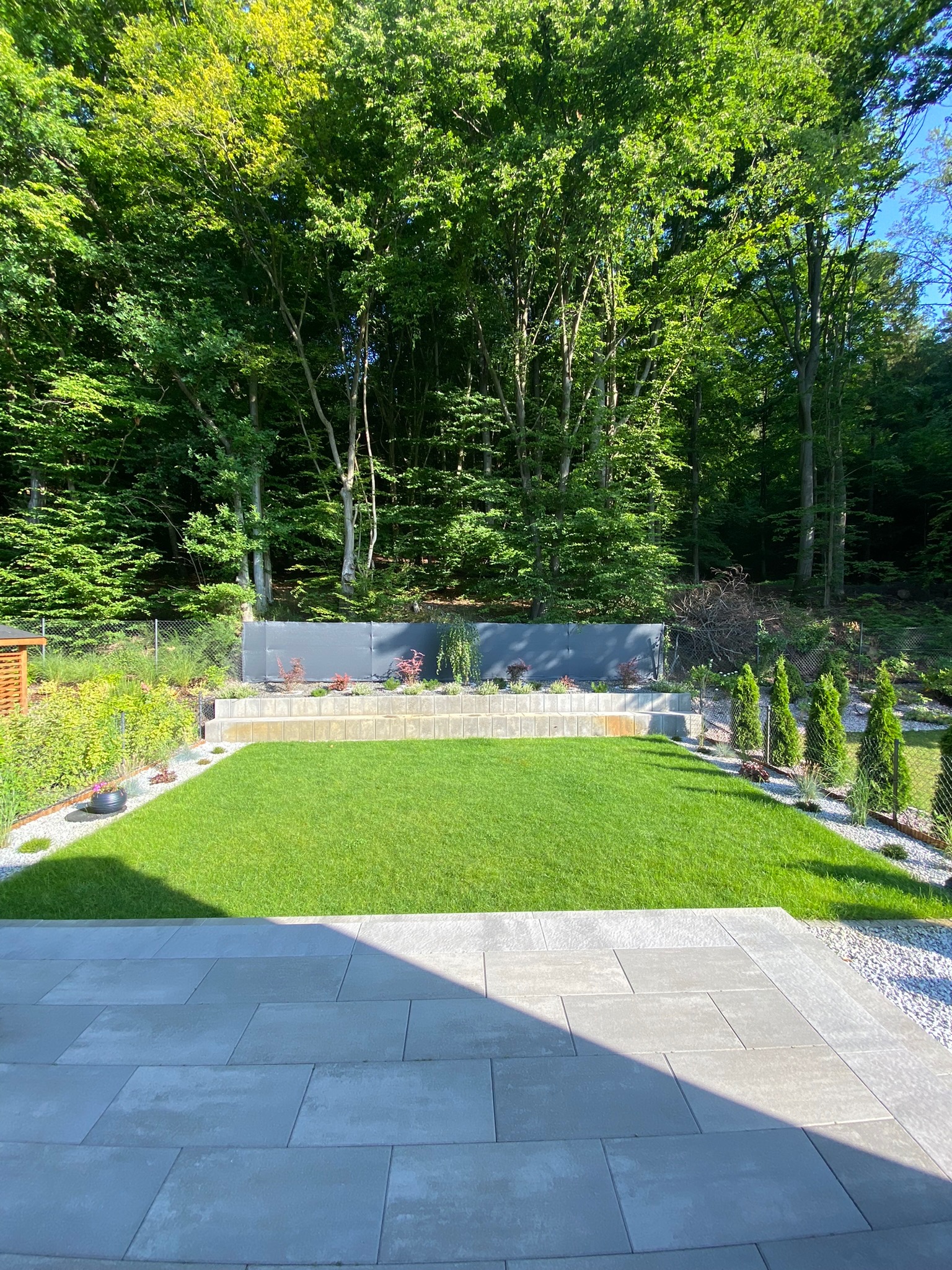 Starannie utrzymany trawnik otoczony rabatami i drzewami, widok z tarasu z szarych płyt.