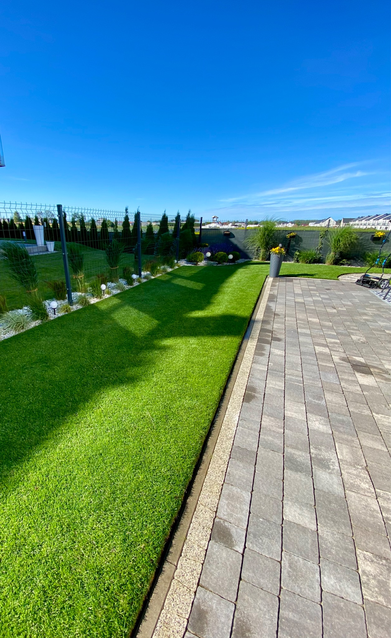 Perfekcyjnie utrzymany trawnik z równo przystrzyżoną trawą graniczący z brukowanym chodnikiem, ozdobne rośliny wzdłuż ogrodzenia, błękitne niebo w tle.