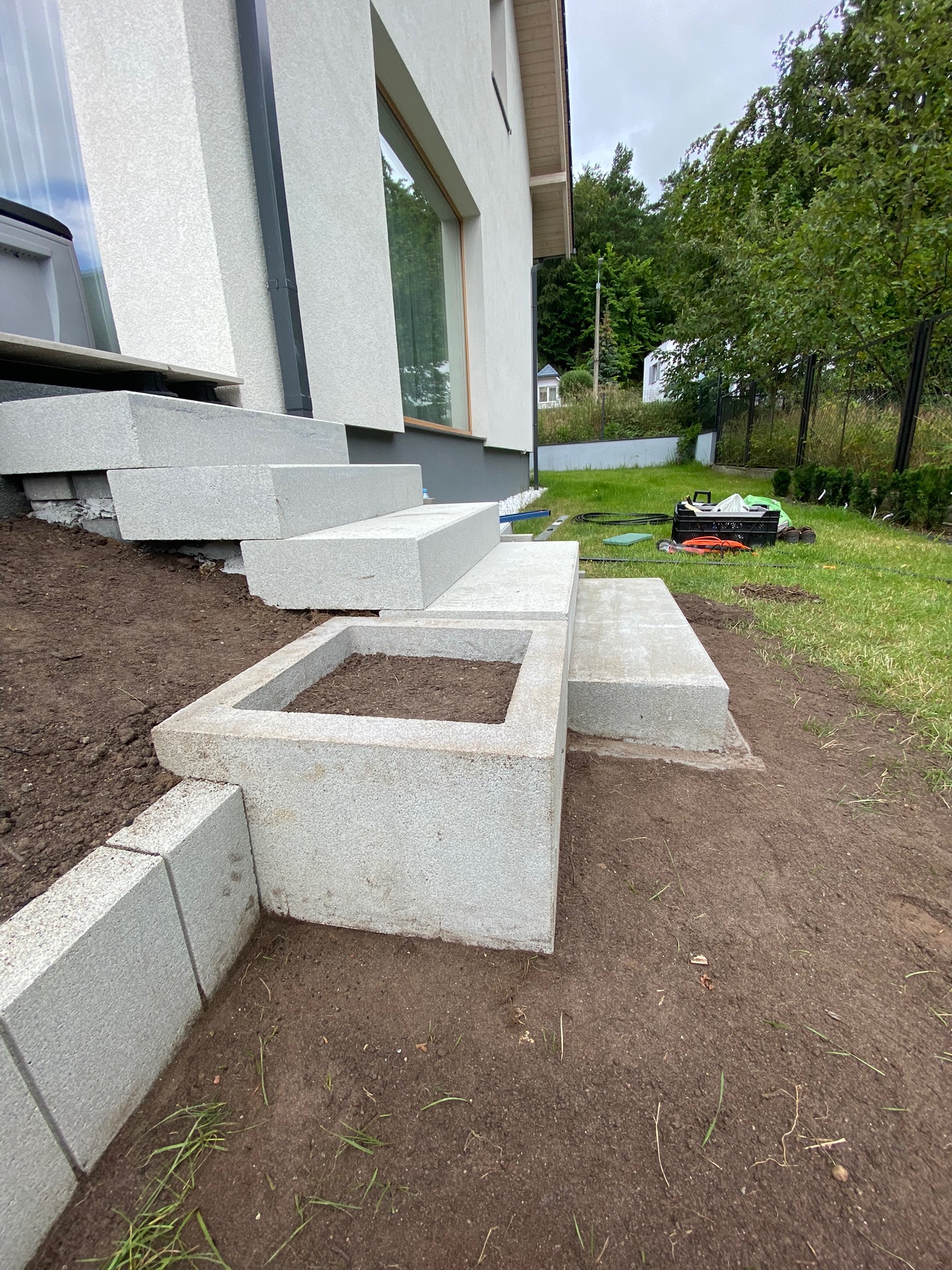 Nowe, granitowe schody wejściowe i obrzeże rabaty z pustaków, z ziemią przygotowaną do sadzenia, na tle trawnika i budynku.